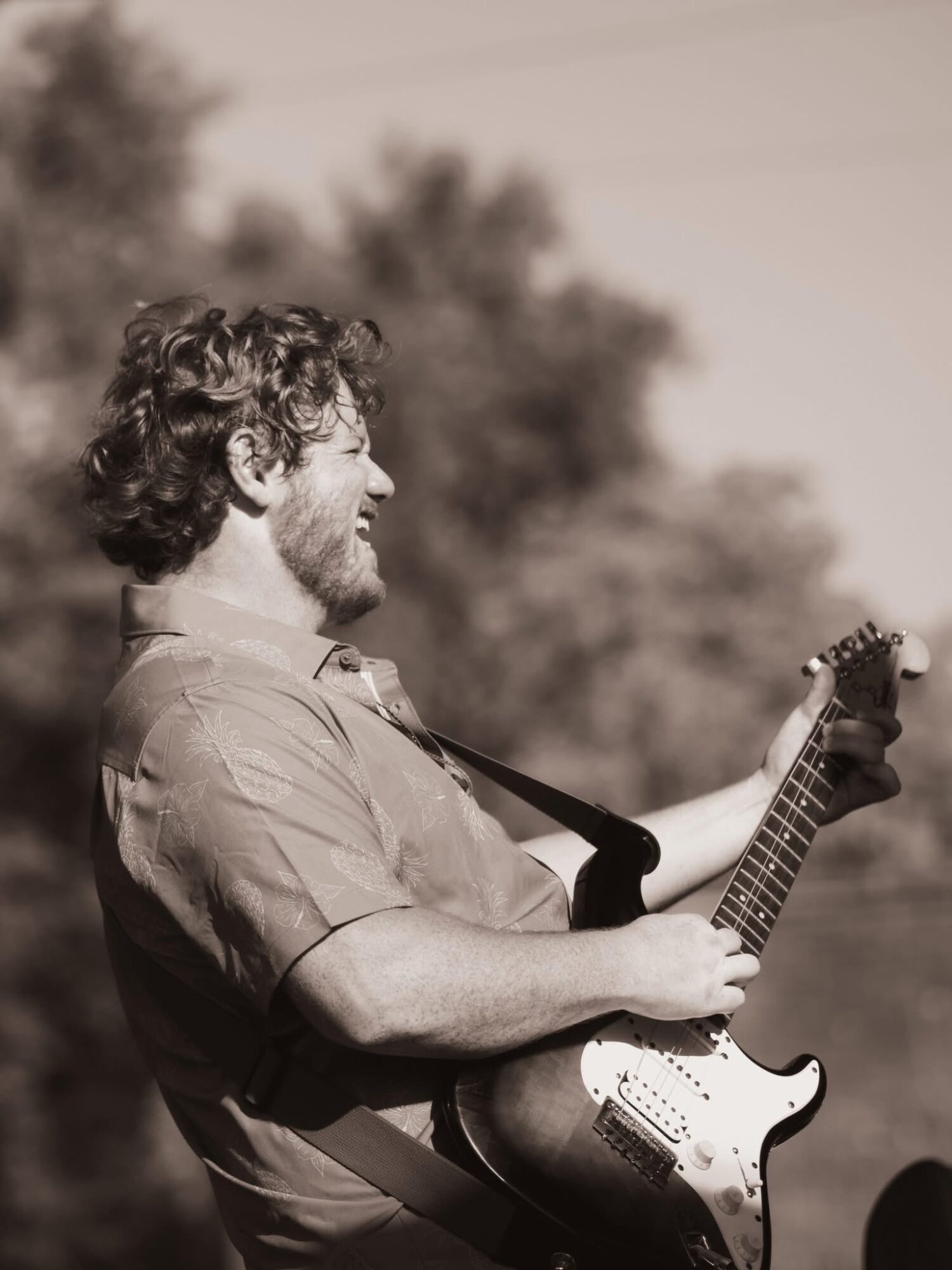 Man with curly hair and beard playing an electric guitar outdoors, smiling, with trees in the background.