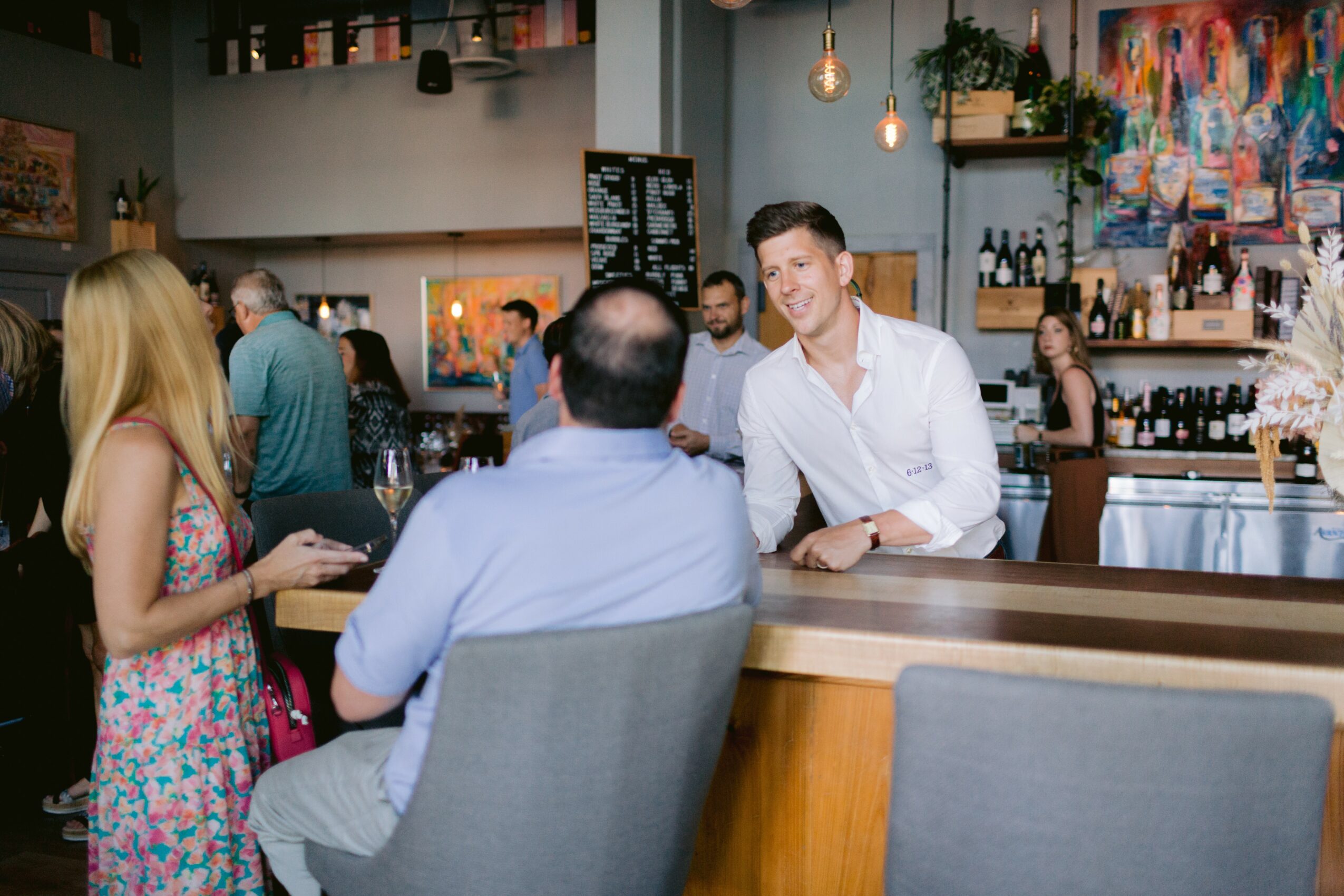 People socializing at a bar with a bartender, colorful artwork, and warm lighting, in a lively indoor setting.