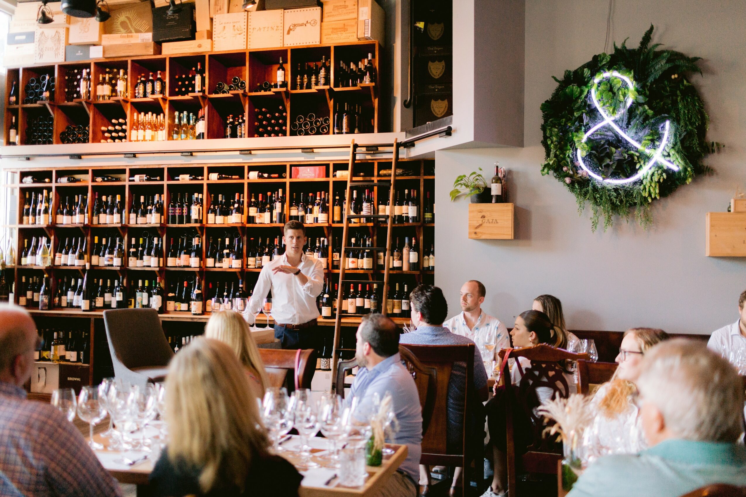 A woman presents to a seated audience in a wine shop with shelves of wine bottles, a wreath on the wall, and tables set for a meal.