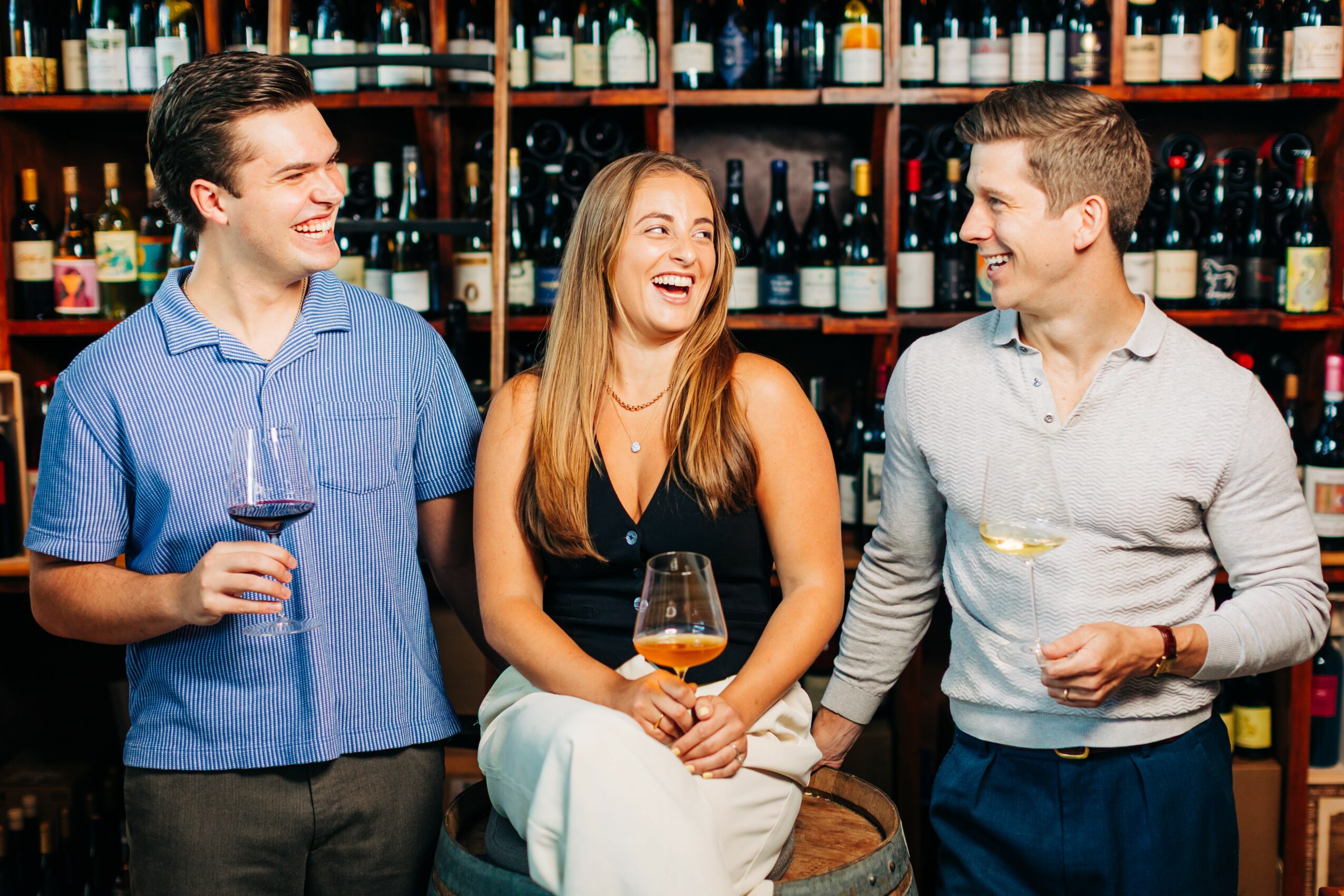 Three people smiling and talking in a wine store, holding glasses of wine, shelves of wine bottles behind them.