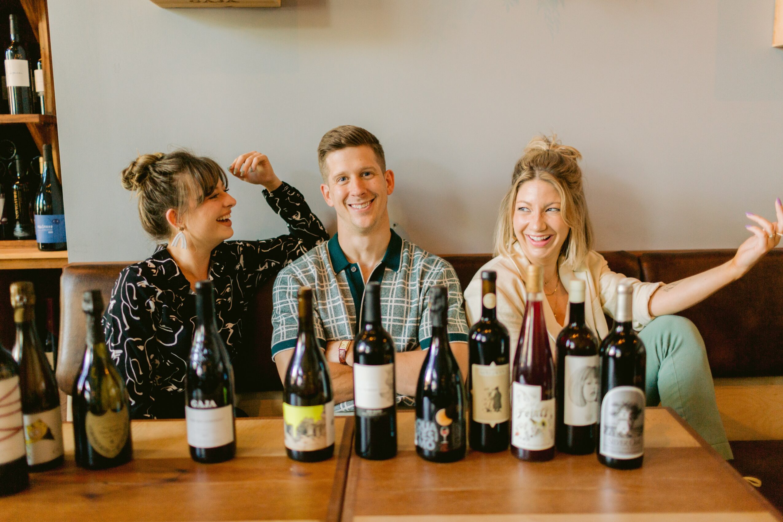 Three people sitting at a table with multiple wine bottles, smiling and engaging with each other.