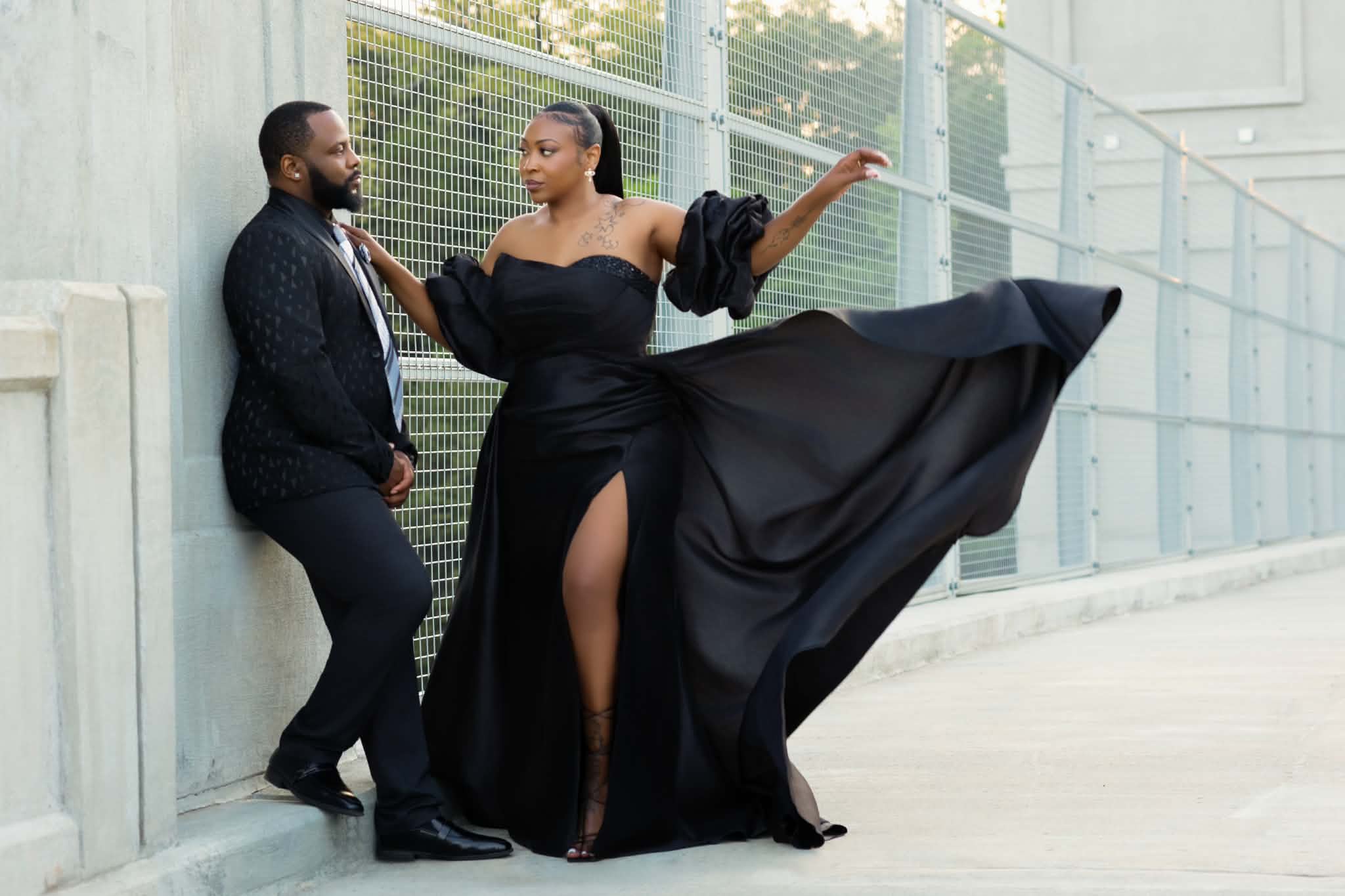 Two people stand outdoors near a metal fence, one in a black dress with a high slit, the other in a dark jacket and pants.