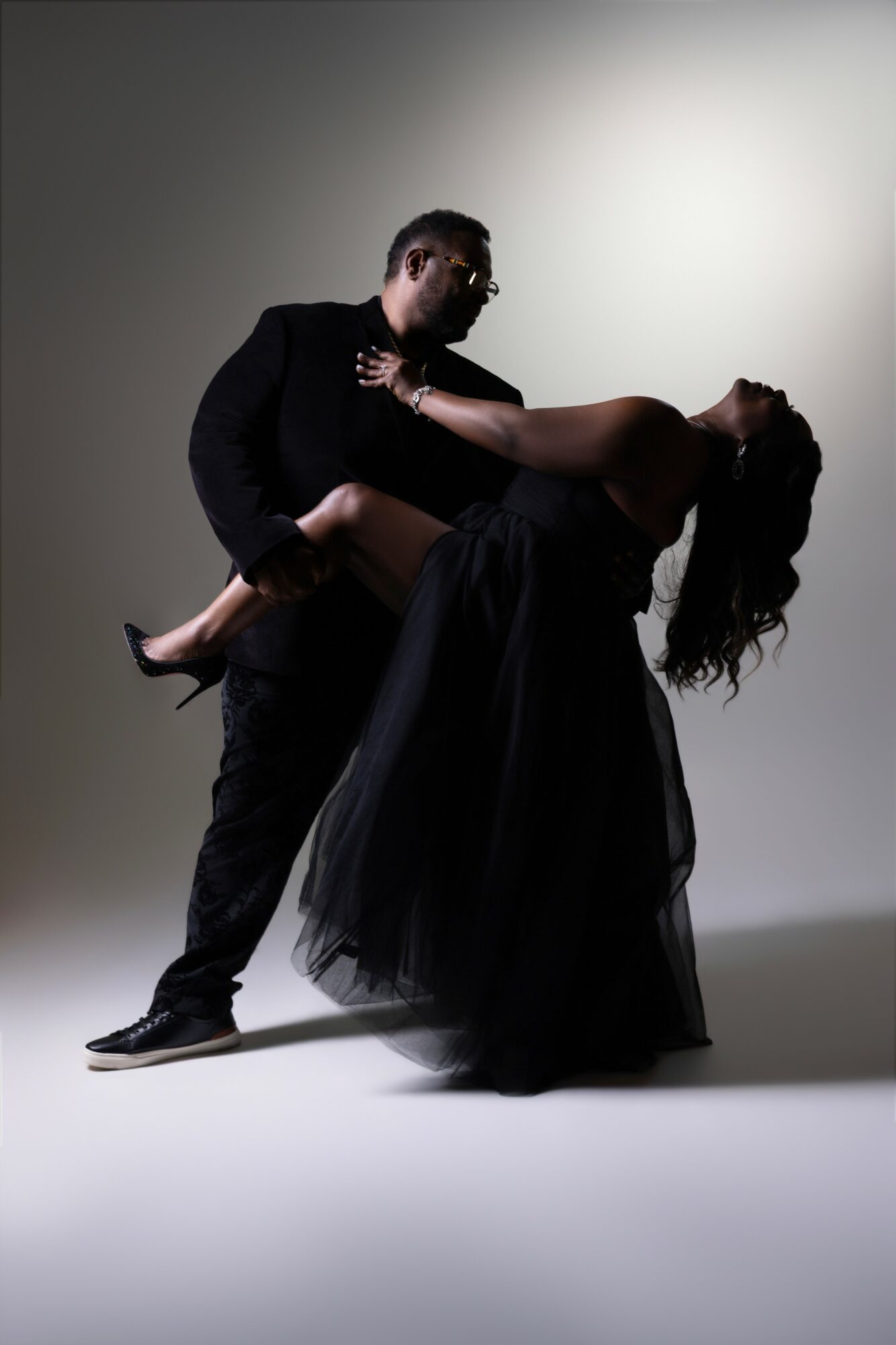 Man in dark suit holding woman in a flowing black dress, dancing against a plain background.