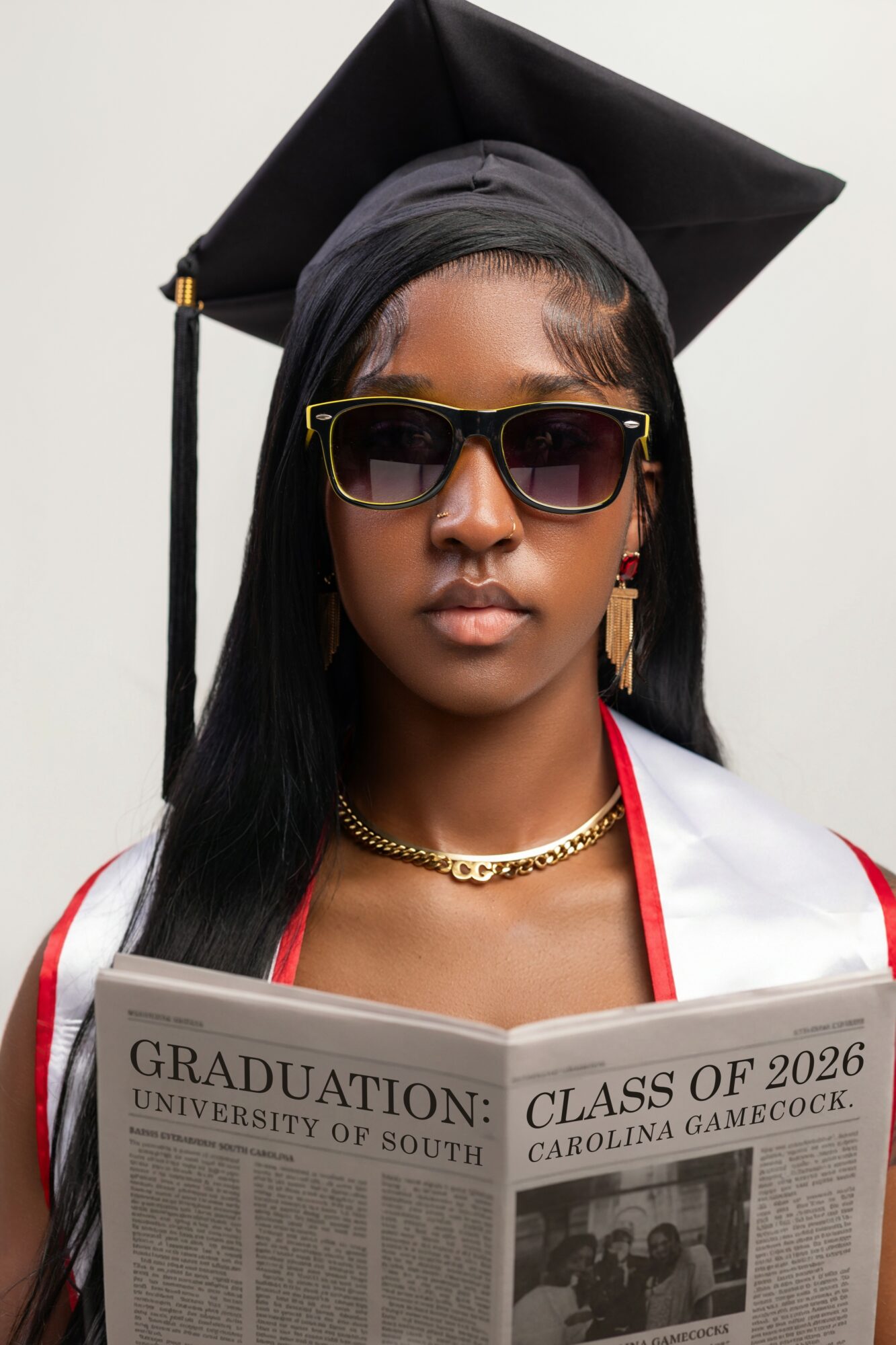 Young woman wearing sunglasses and graduation cap, holding a newspaper, with a white background.