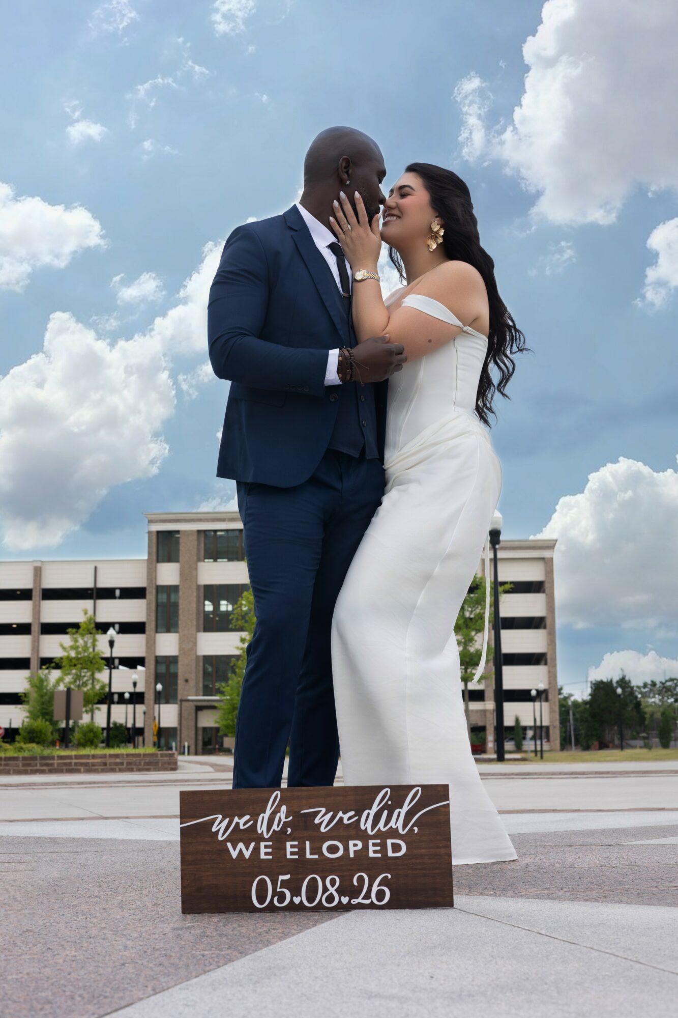 A couple in wedding attire embracing outdoors with a building and cloudy sky in background.