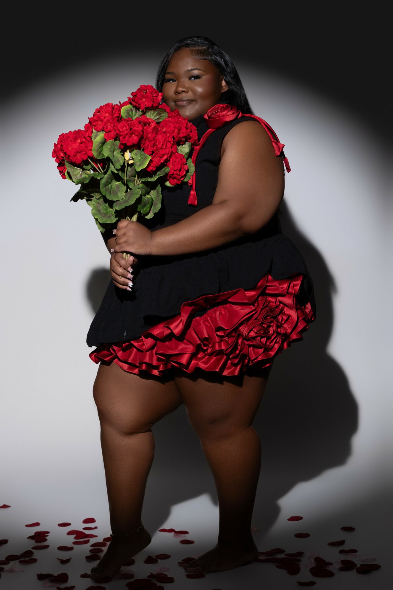 Woman holding a large bouquet of red flowers, wearing a black and red ruffled dress, standing on a white background with flower petals on the floor.