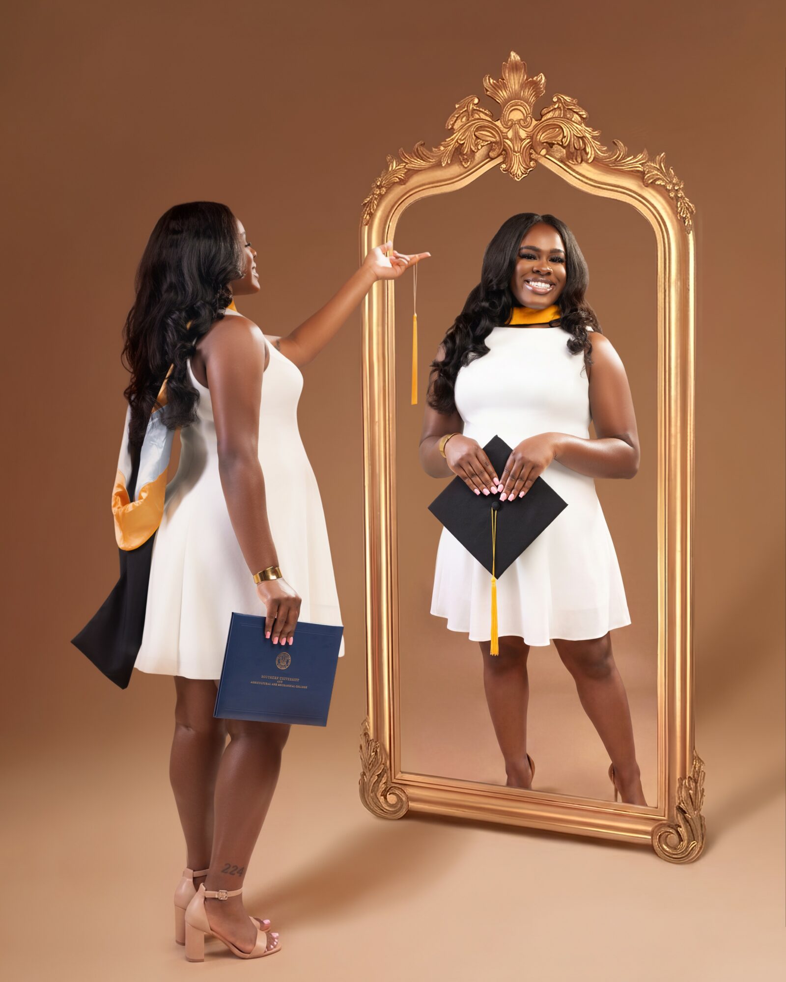 Woman in a white dress looking at her reflection in a large ornate mirror, holding a diploma, smiling.