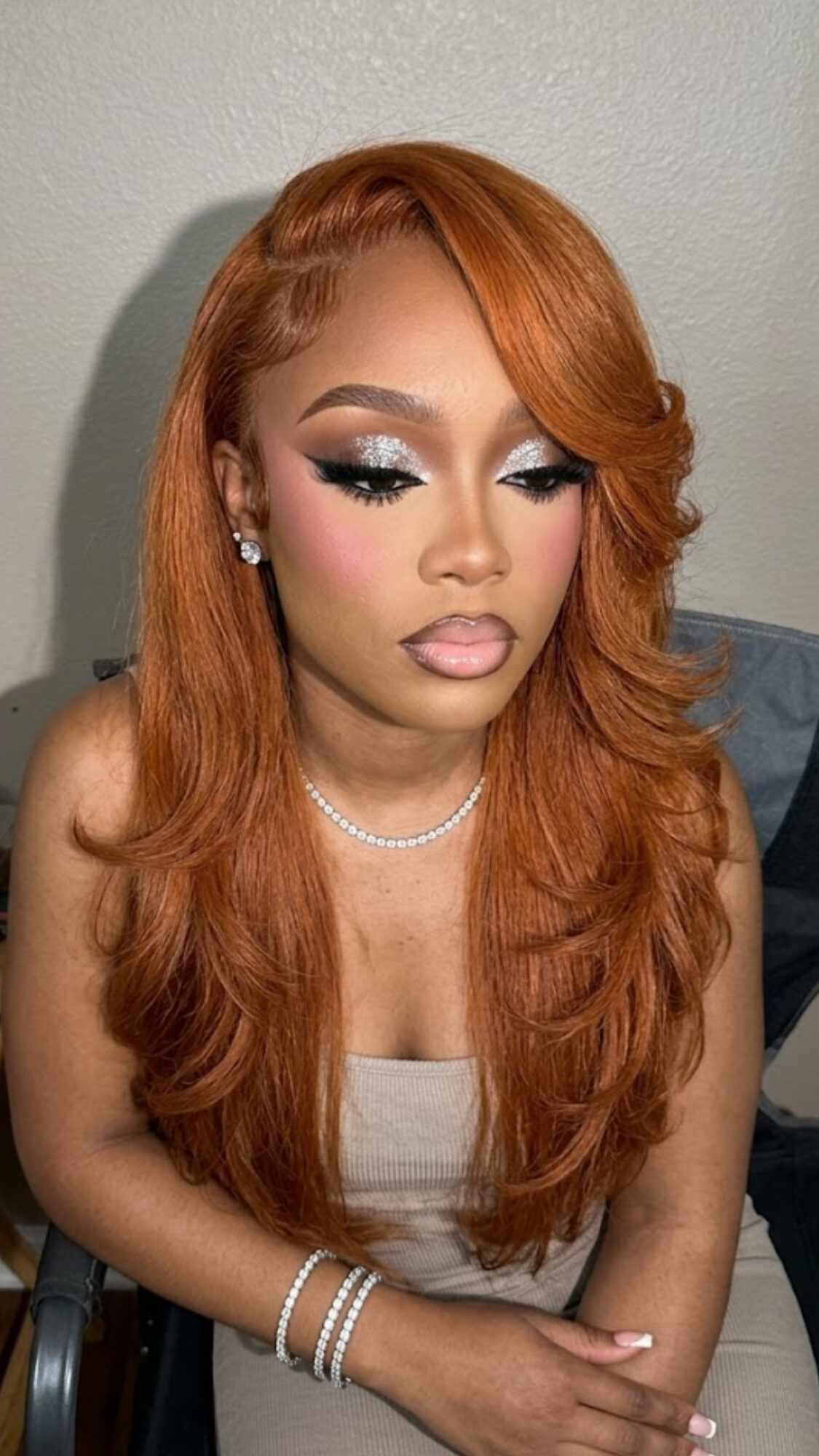 Woman with long, wavy orange hair and makeup, wearing earrings, a necklace, and a sleeveless top, sitting against a plain wall.