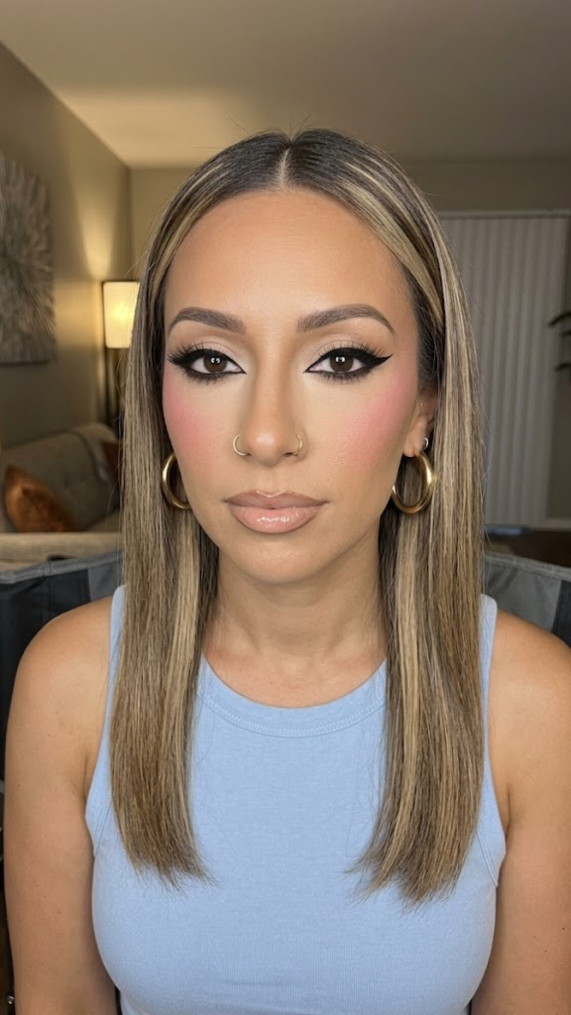 Young woman with straight, shoulder-length hair, makeup, and gold hoop earrings, wearing a light blue top indoors.