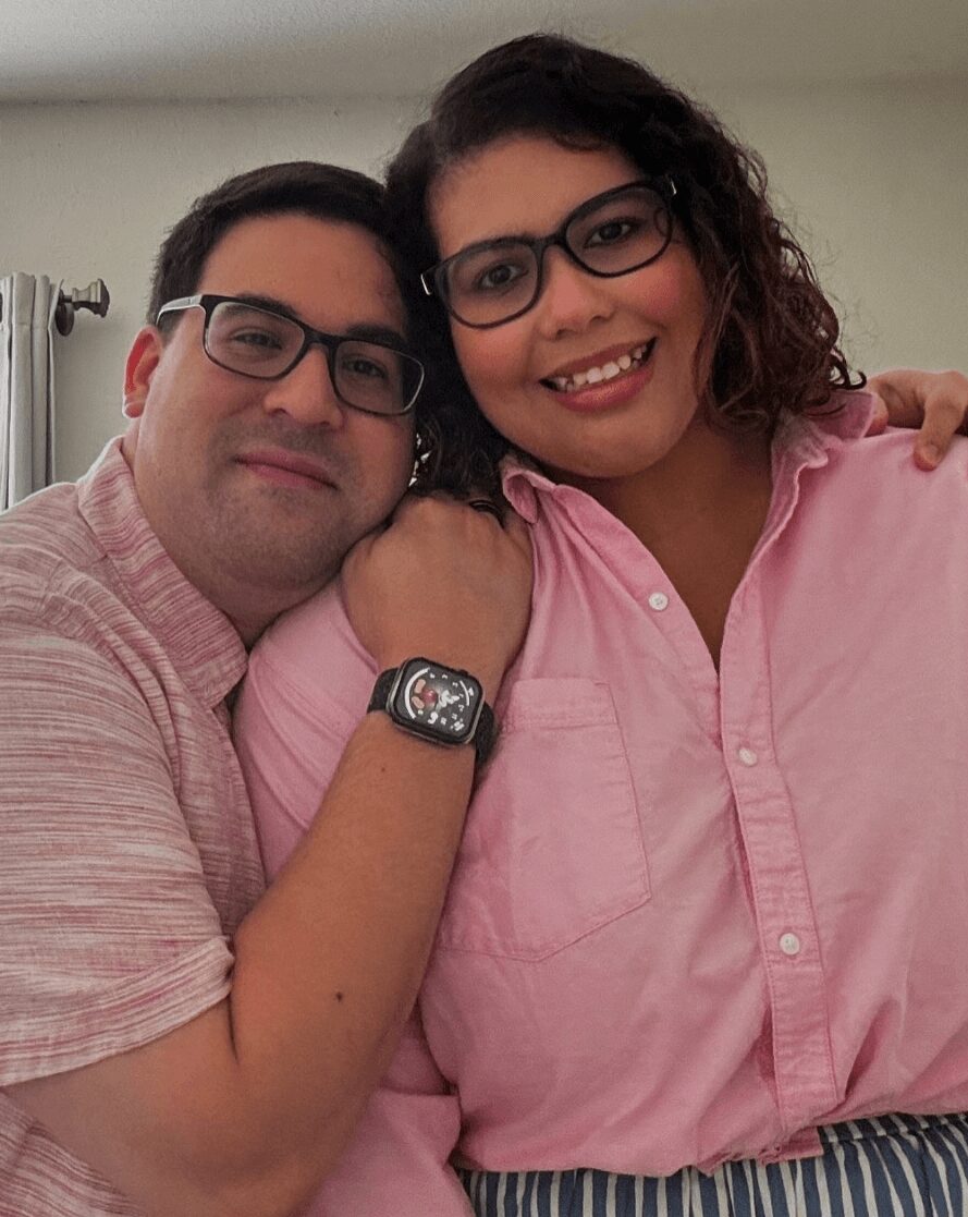 A man and woman with glasses smiling, close together, woman wearing a pink shirt, man in a light pink shirt, indoors.