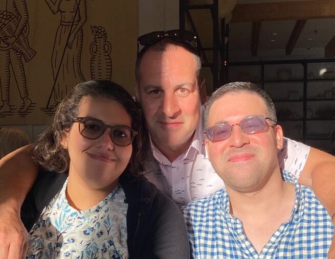 Three people smiling, two wearing glasses, in an indoor setting with wall art and shelves in the background.