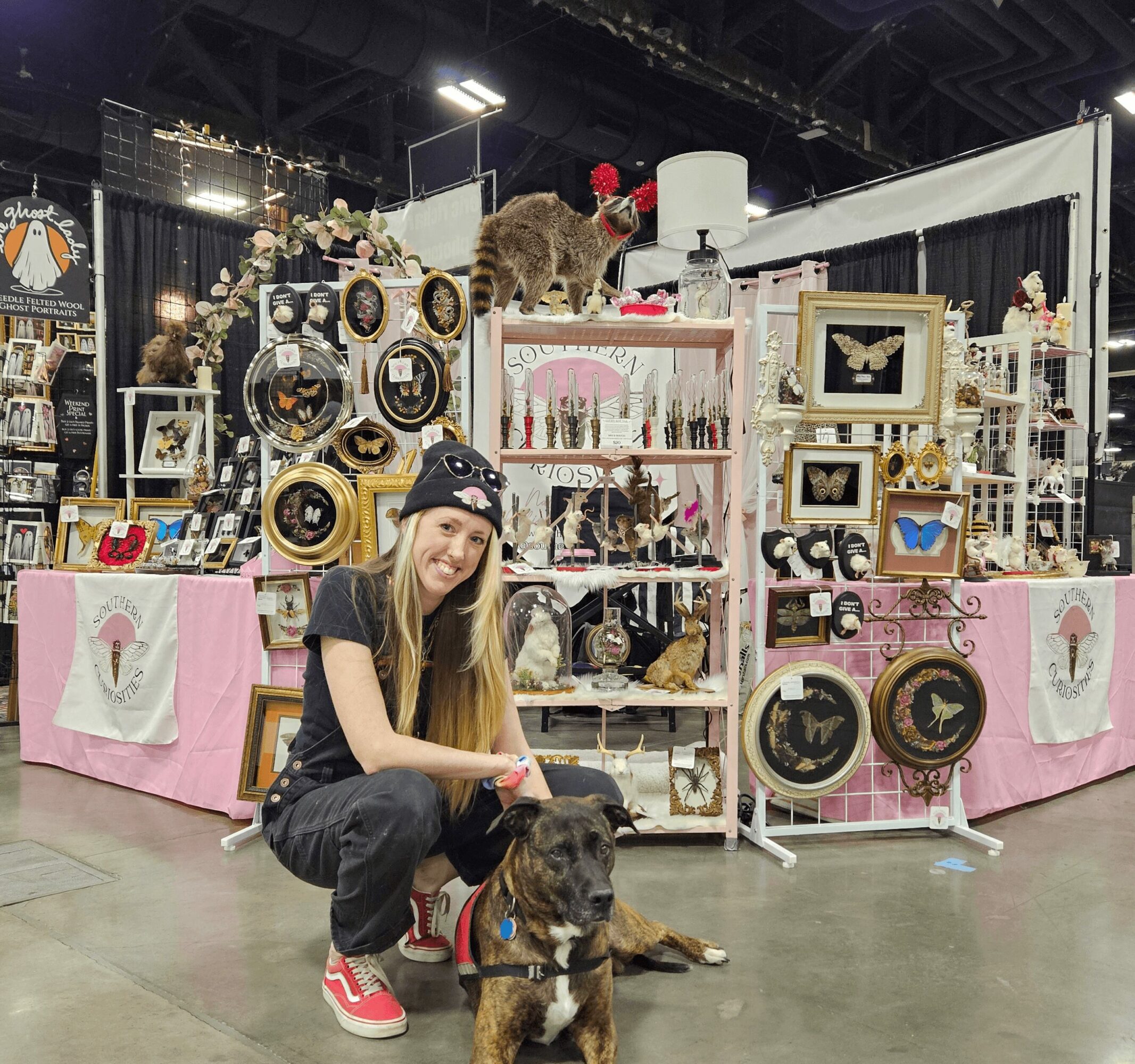 Young woman with a dog in front of a jewelry display at an indoor market, smiling at the camera.