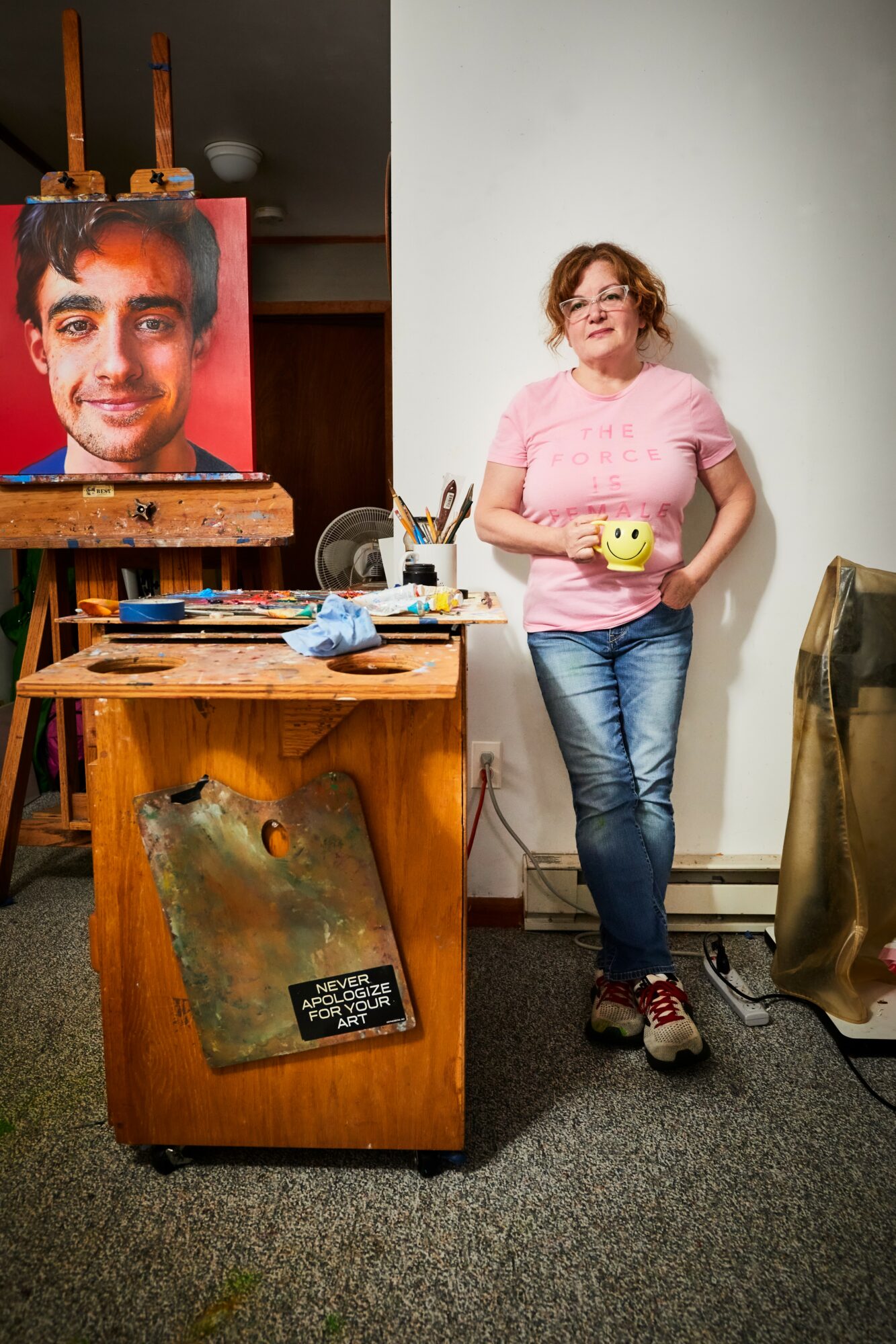 Woman with curly hair in pink shirt and jeans holding a yellow smiley face mug, standing next to an art easel with a portrait, in a room with art supplies and a wooden cabinet.