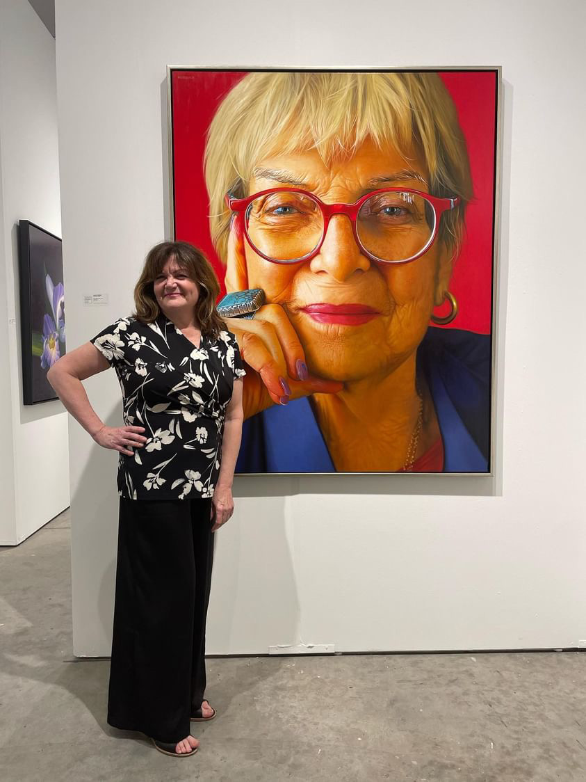 Woman standing in art gallery, large portrait of an older woman with glasses behind her, white walls, concrete floor.