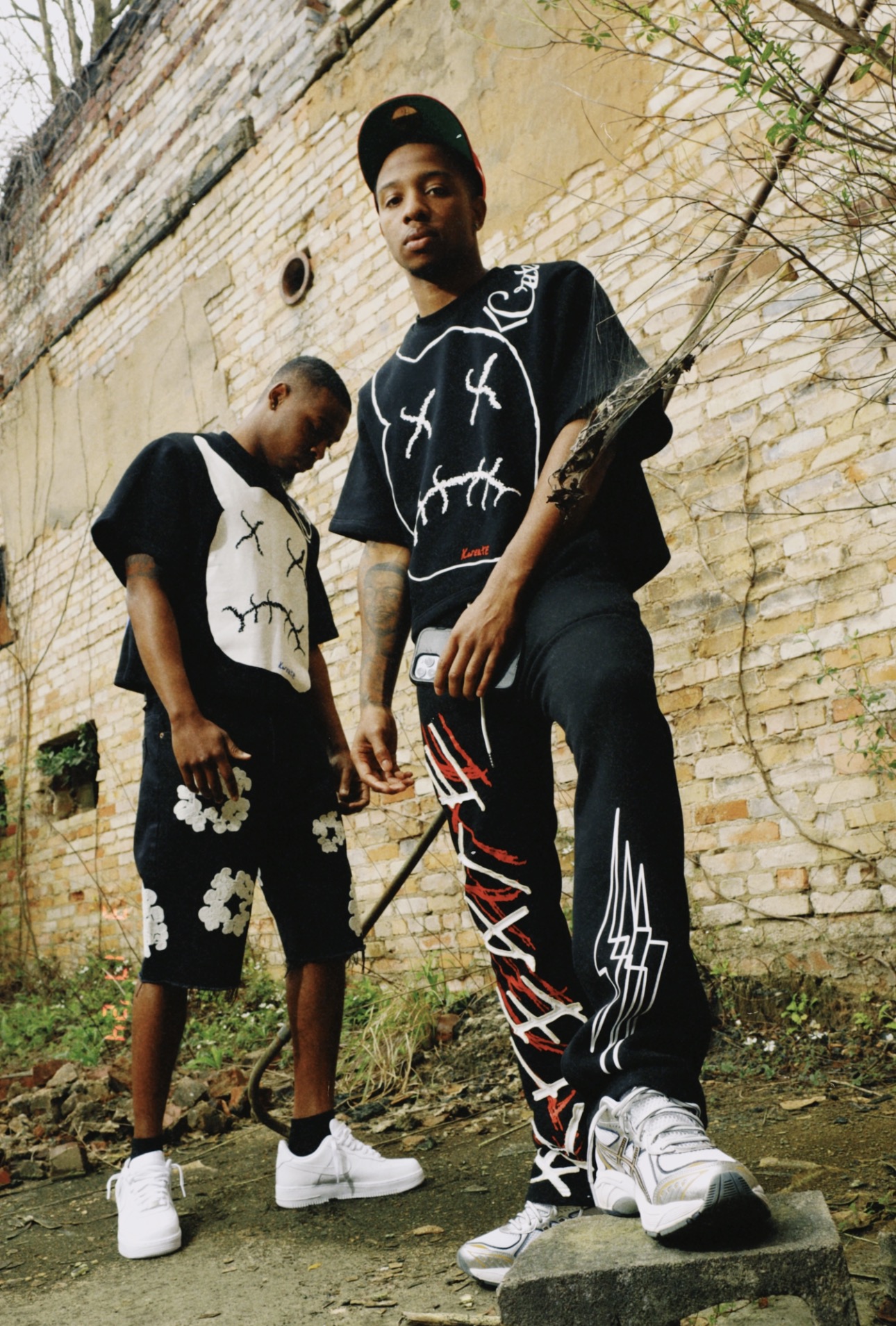 Two young men standing outdoors against a brick wall, one on a skateboard, both wearing black clothing with white and red designs.