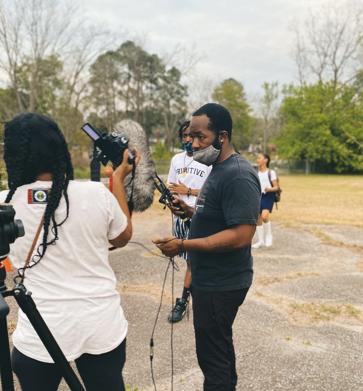 Group of people outdoors, some wearing masks, one person holding a camera, others with microphones, trees in background.
