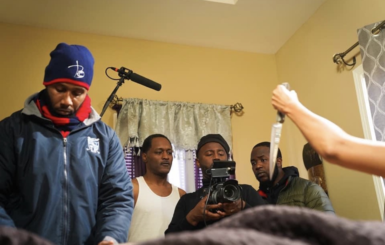 Four men standing in a room with a camera, one holding a camera, others looking down or at the camera, yellow walls, window with curtain.