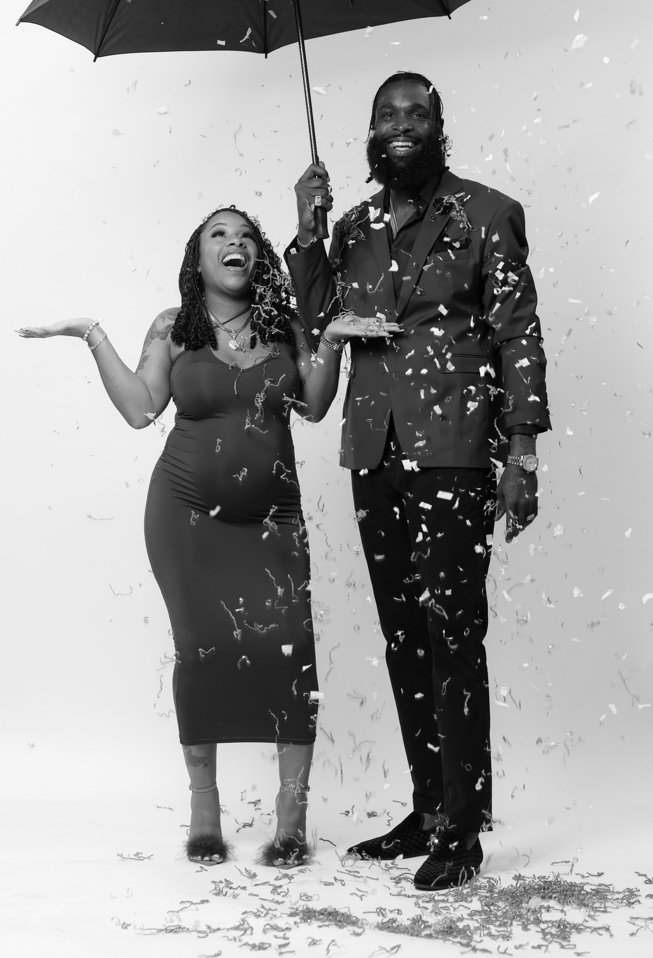 A smiling woman and man stand together under an umbrella, surrounded by falling snow or confetti, with a plain background.
