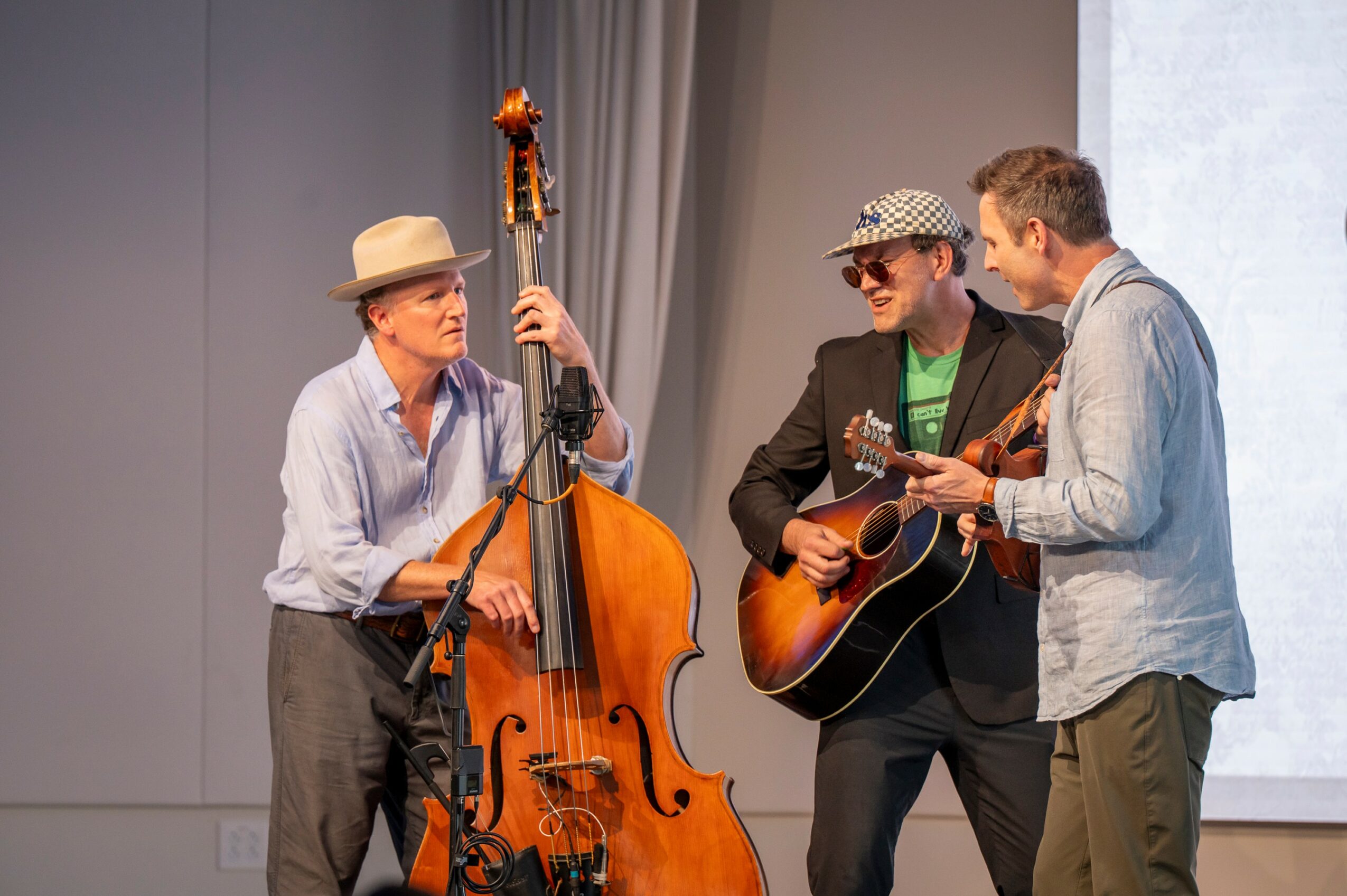 Three men performing music on stage, one with a double bass, another with a guitar, and the third with a small instrument, all engaged in a musical performance.