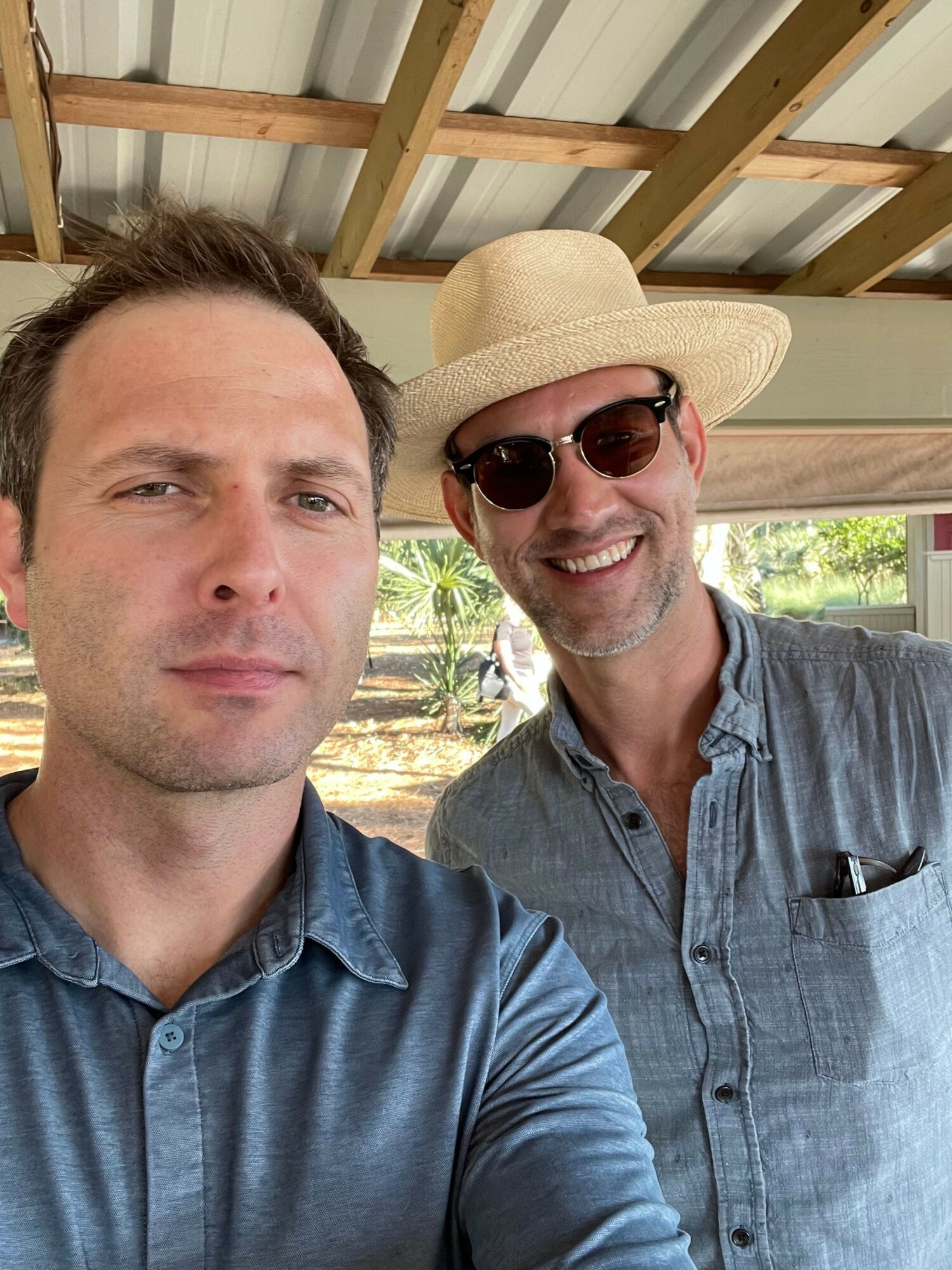 Two men outdoors, one wearing a straw hat and sunglasses, the other with short hair, both smiling.