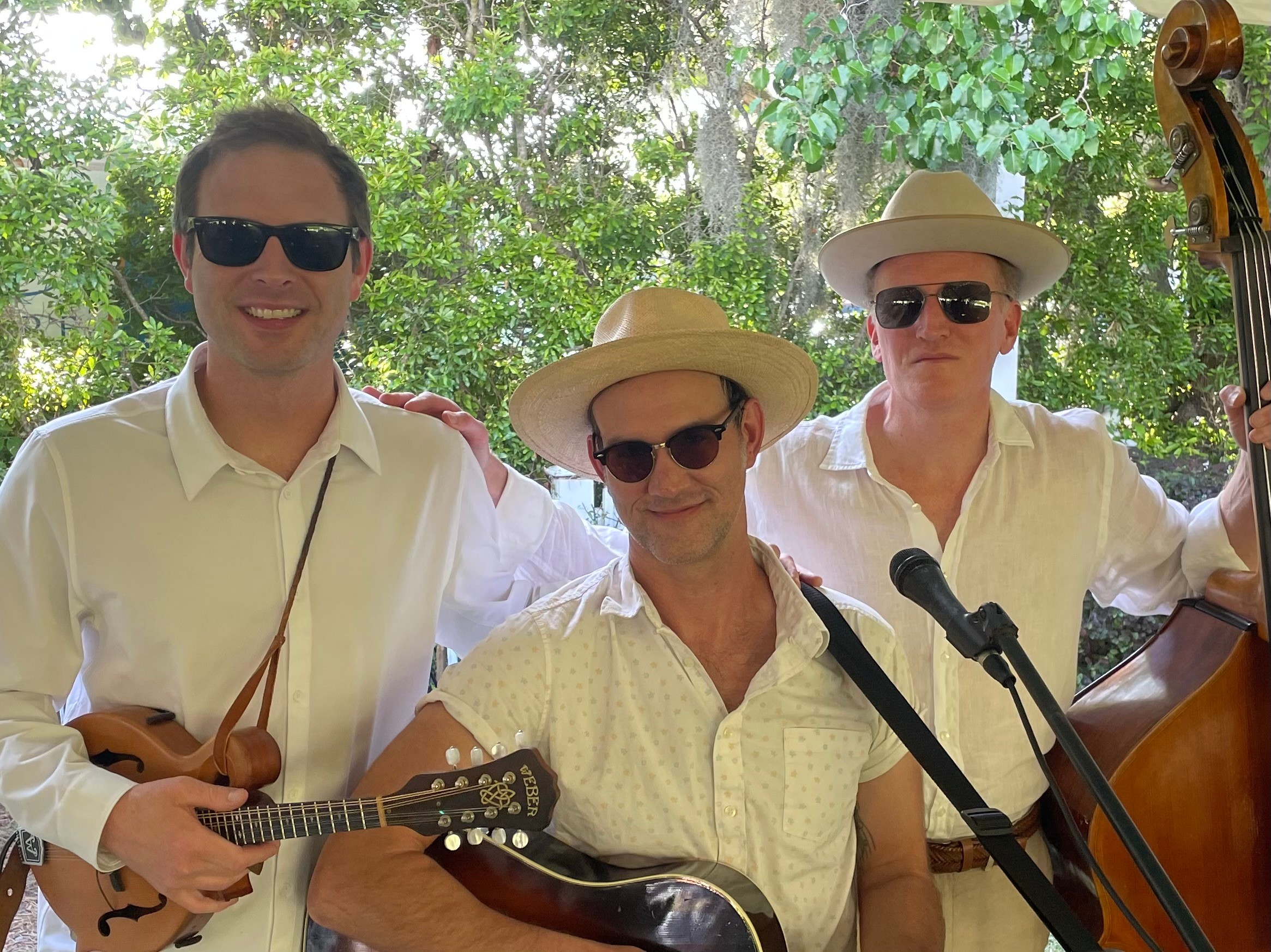 Three men wearing sunglasses and hats, holding musical instruments outdoors with trees in the background.