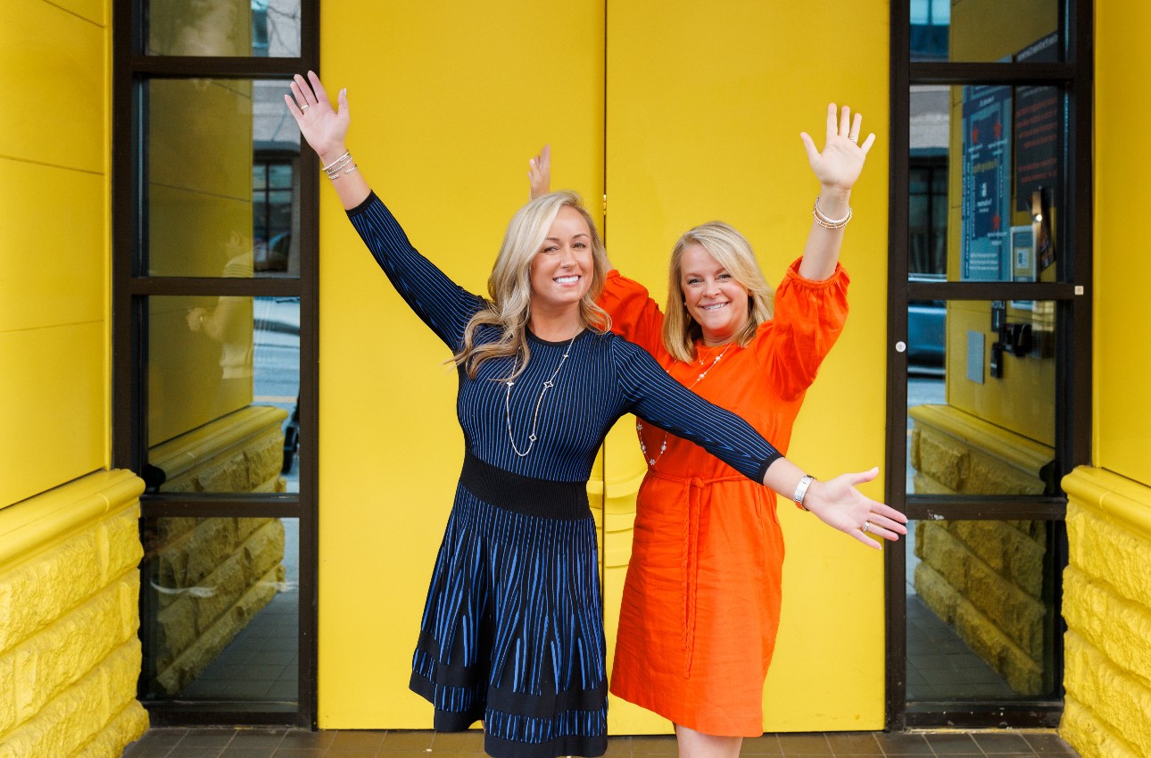 Two women with blonde hair smiling and waving, standing in front of a yellow wall with black-framed glass doors.