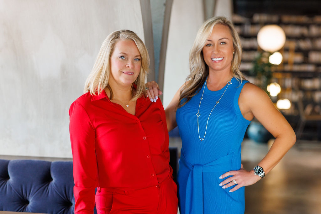 Two women standing indoors, one in a red dress and the other in a blue dress, smiling. Background includes a bookshelf and lights.