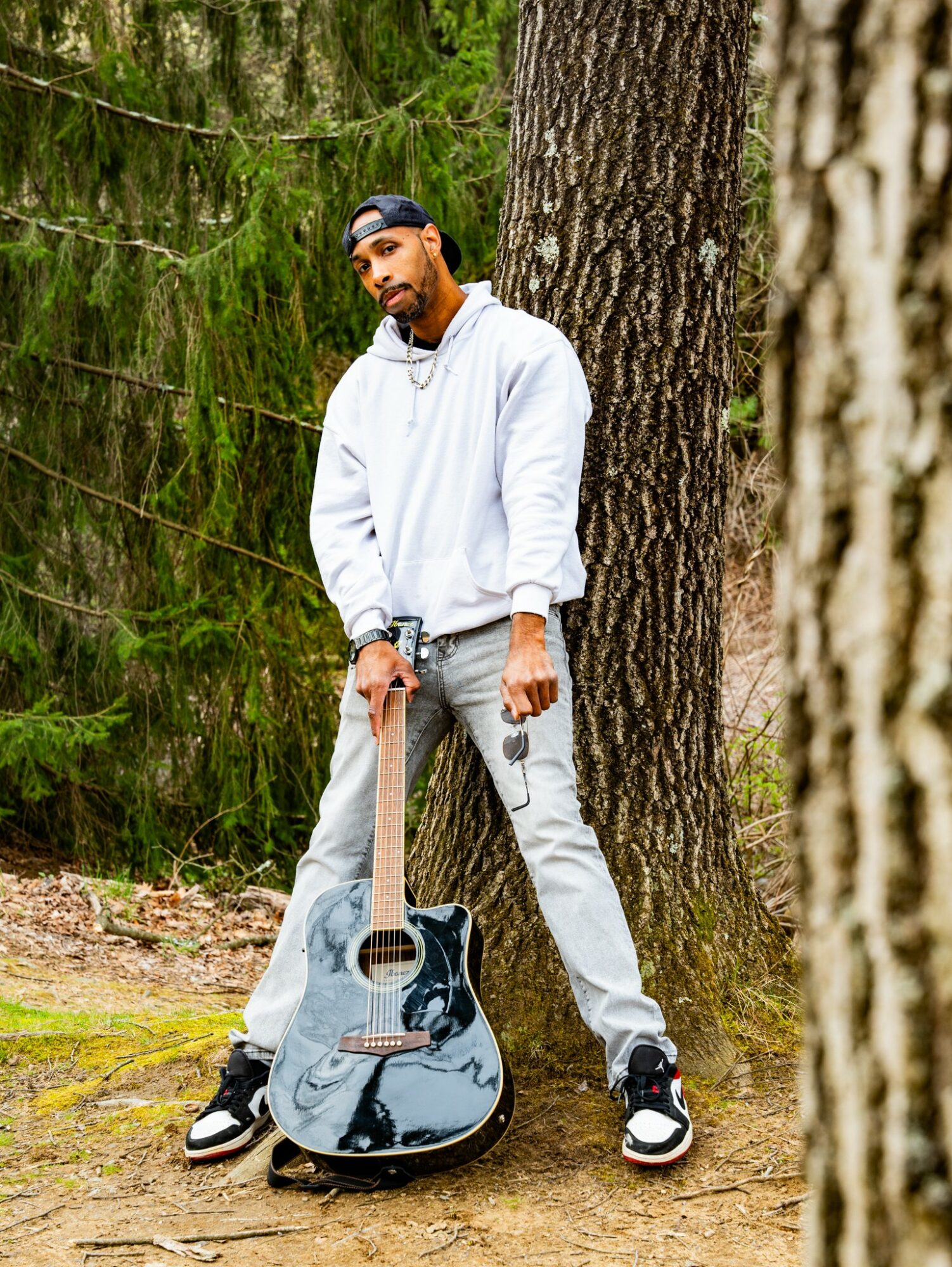 Young man with a beard in a white hoodie and gray pants stands outdoors next to a large tree, holding a guitar case.