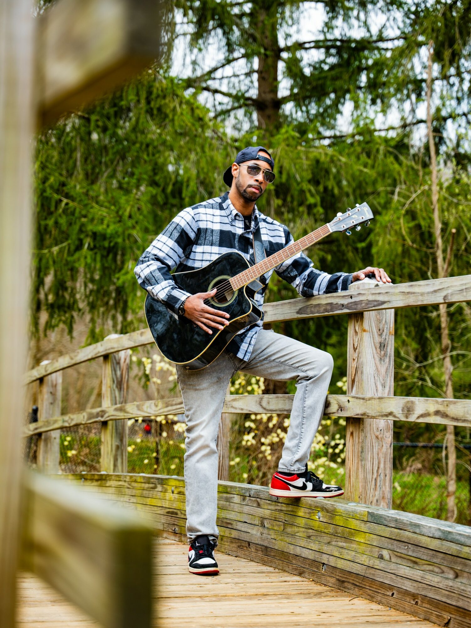 Man with sunglasses and beard playing guitar on wooden bridge in forest, wearing plaid shirt, gray pants, and red sneakers.