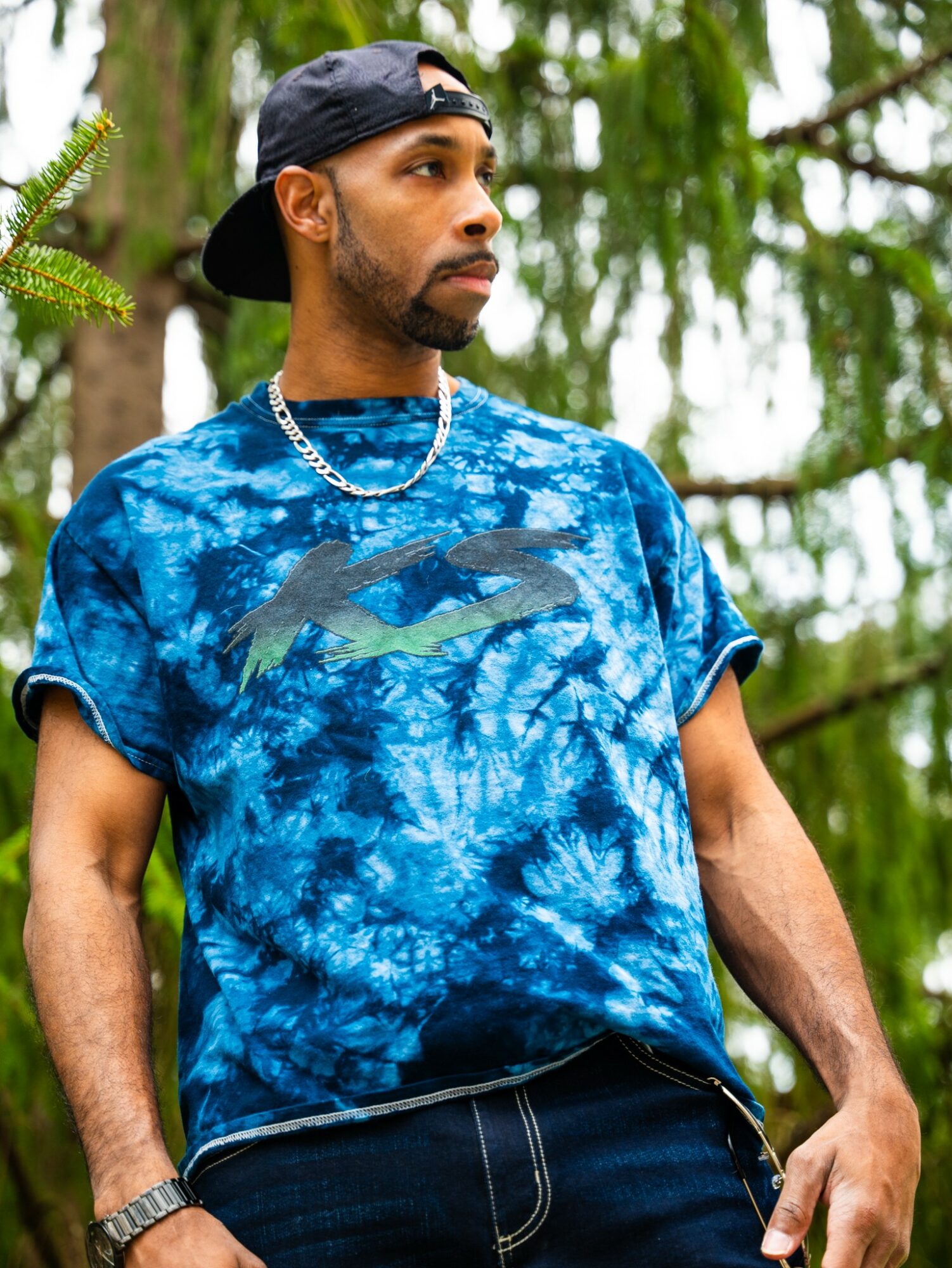 Man wearing a blue tie-dye shirt, black cap backwards, and silver chain, standing outdoors with trees in background.