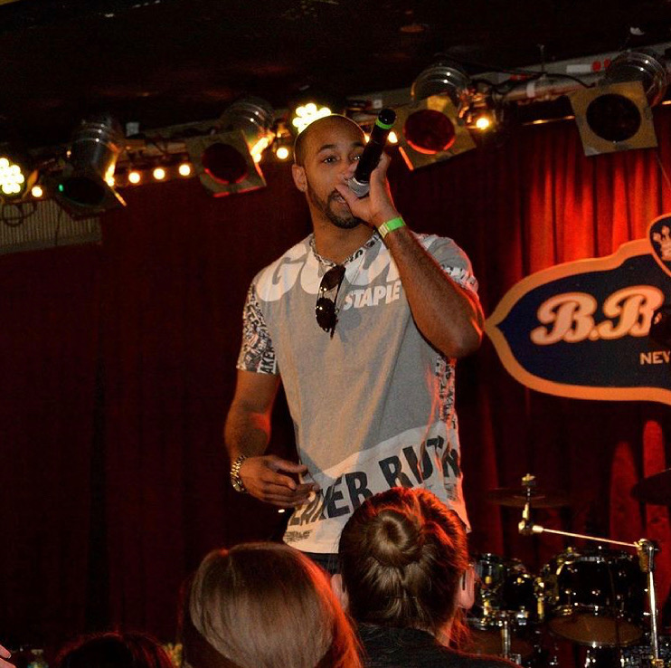 Man holding microphone on stage with red curtain background, audience in foreground, stage lights above, and a drum set nearby.