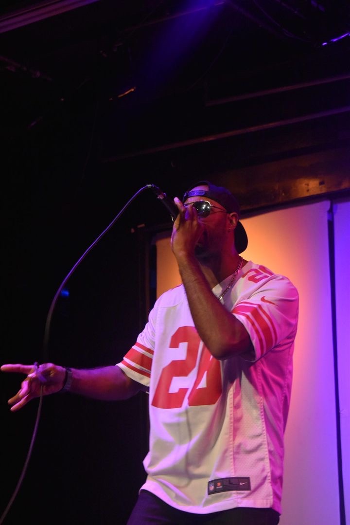 Person singing into microphone on stage, wearing a white sports jersey with red accents and the number 22, with colorful lighting