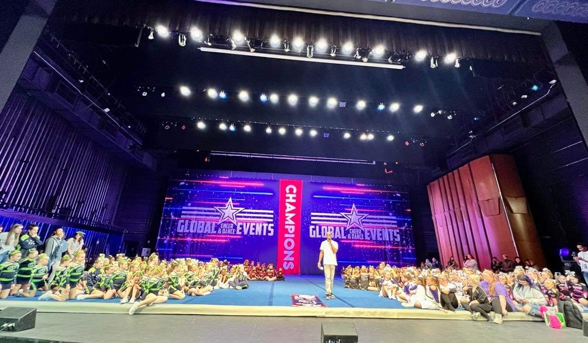 Stage with large screen displaying 'Global Events' and 'Champions,' surrounded by flowers and people, in an auditorium.