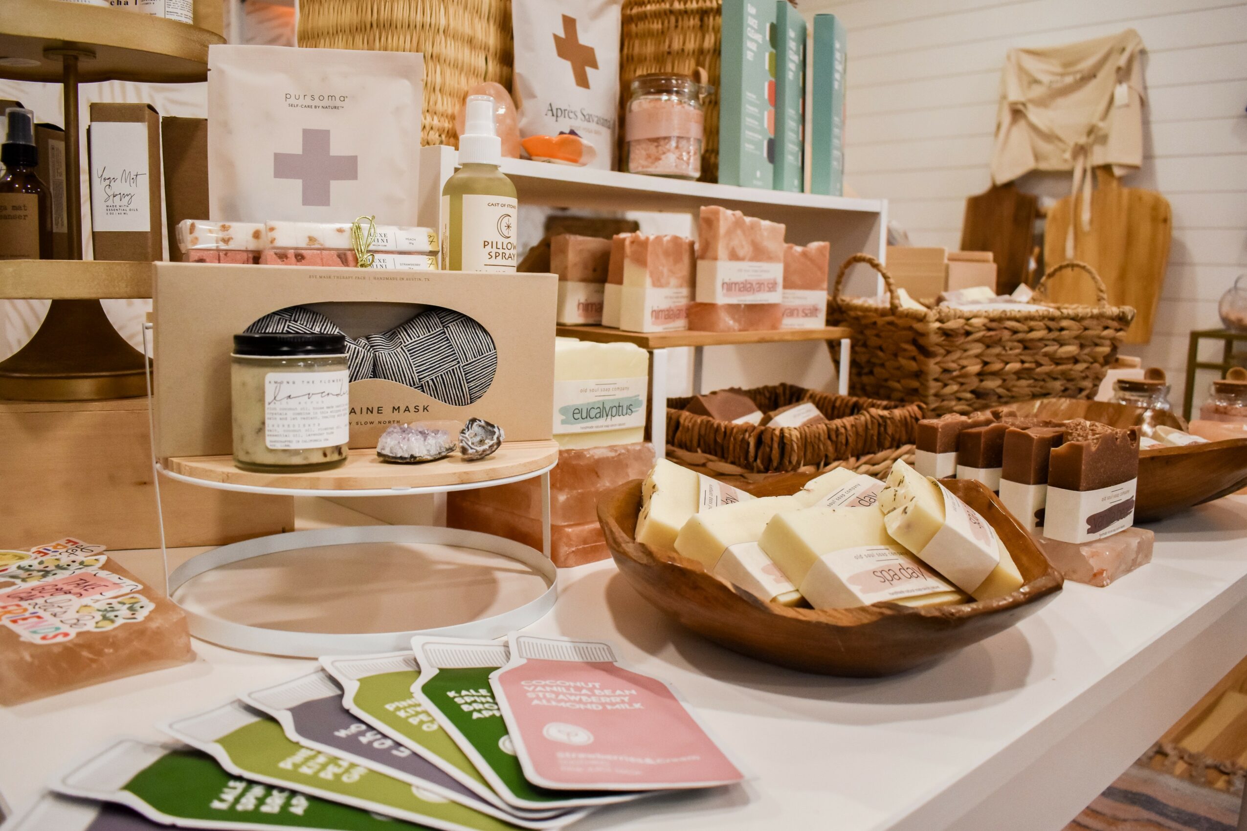 Display of soap bars, skincare products, and informational cards on a white table in a store, with shelves and baskets in background.