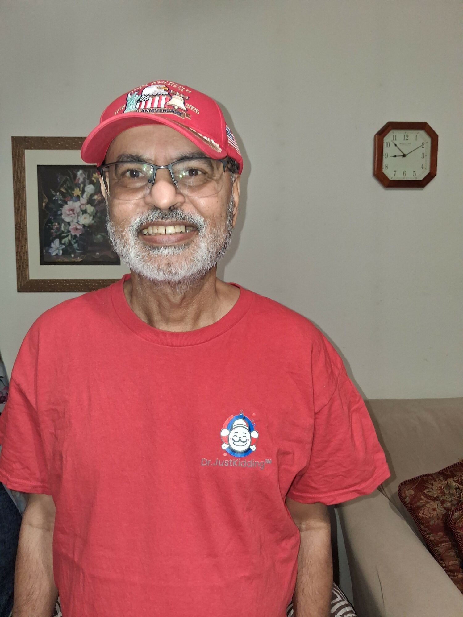 Older man with glasses, beard, and a red cap, smiling, wearing a red shirt, indoors with a clock and picture behind him.