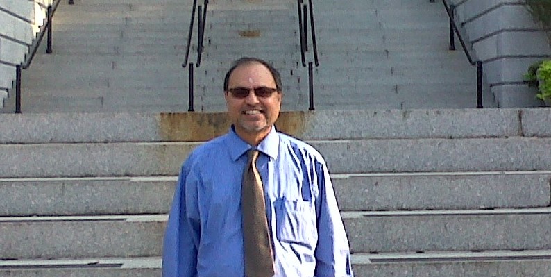 Man in blue shirt and tie standing on stairs outdoors, smiling, wearing sunglasses.