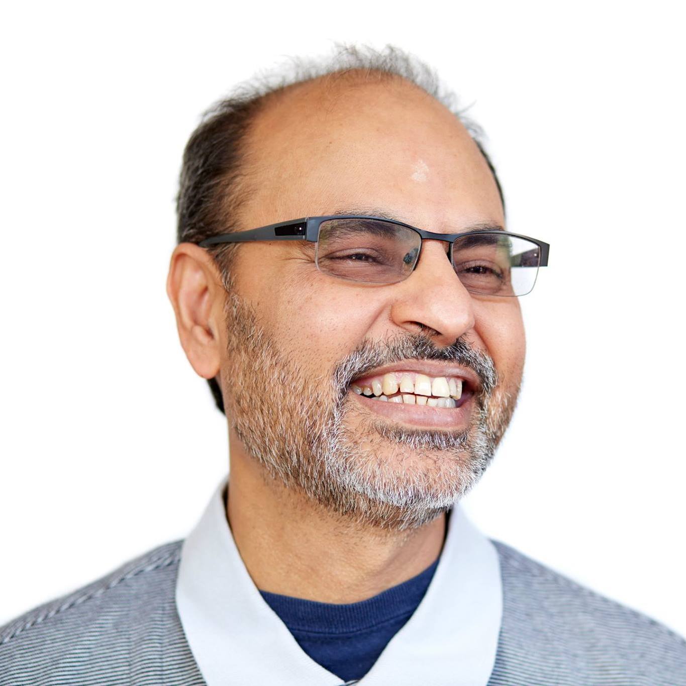 Smiling man with glasses, beard, and gray hair, wearing a light gray sweater and white shirt, against a white background.