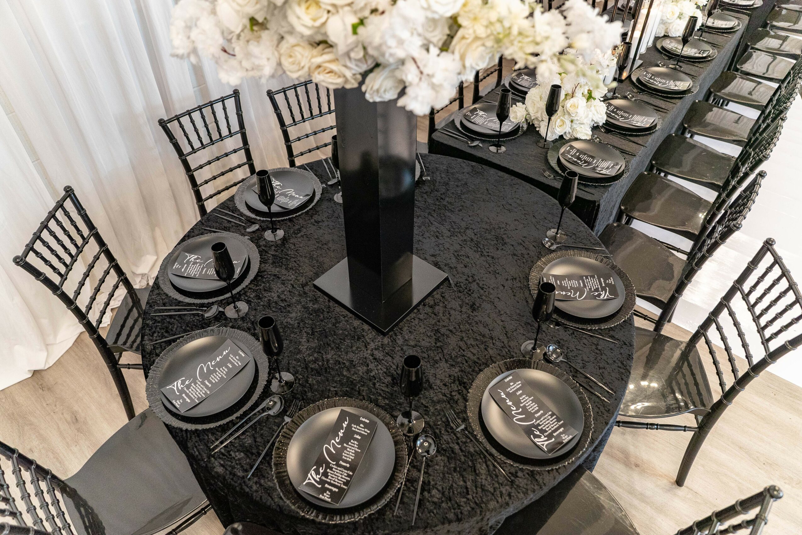 Elegant black dining table with black plates, silverware, and a large floral centerpiece, surrounded by black chairs with vertical slats.