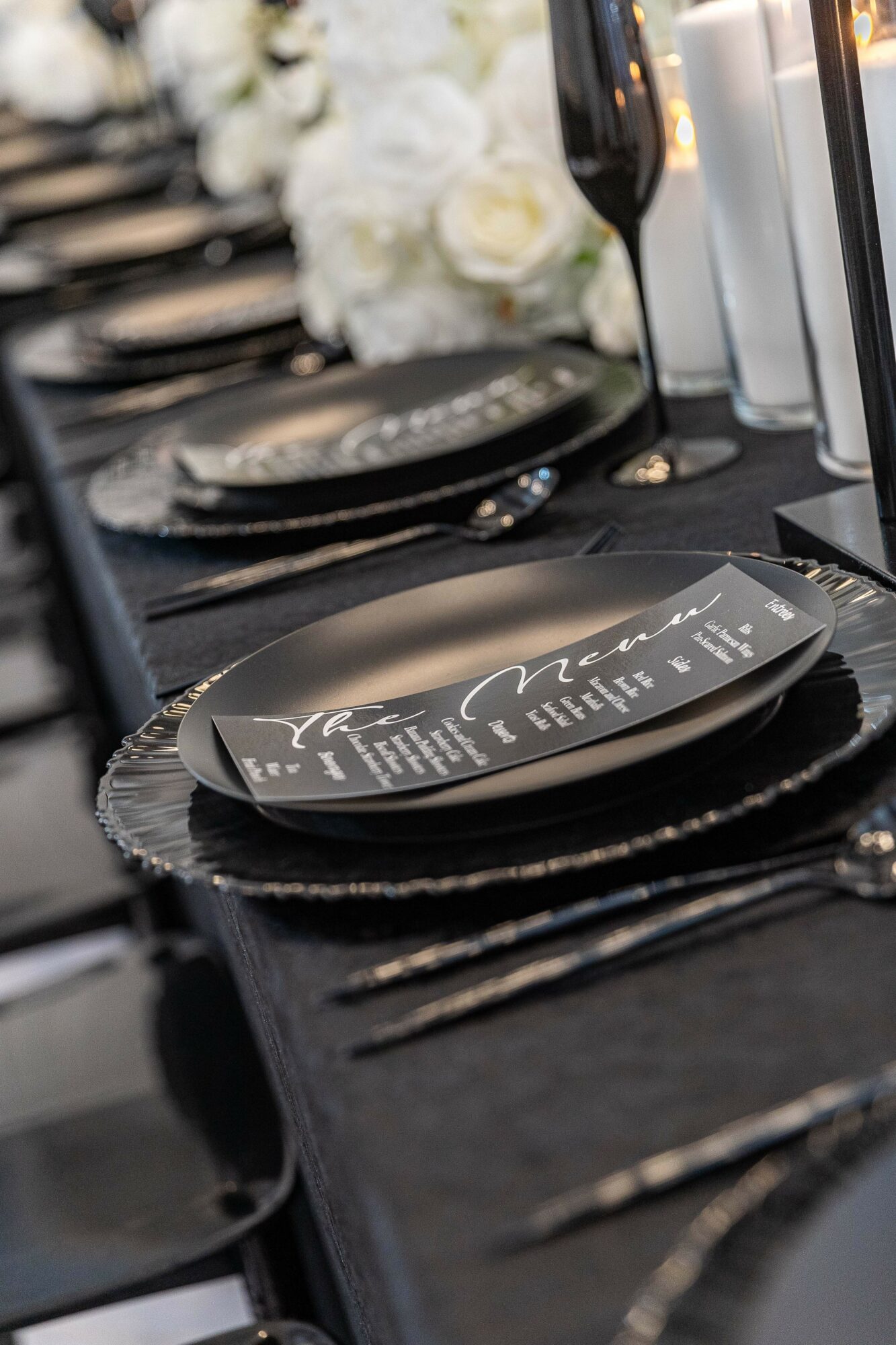 Elegant table setting with black plates, silverware, a menu, and a glass of red wine, decorated with white flowers in the background.