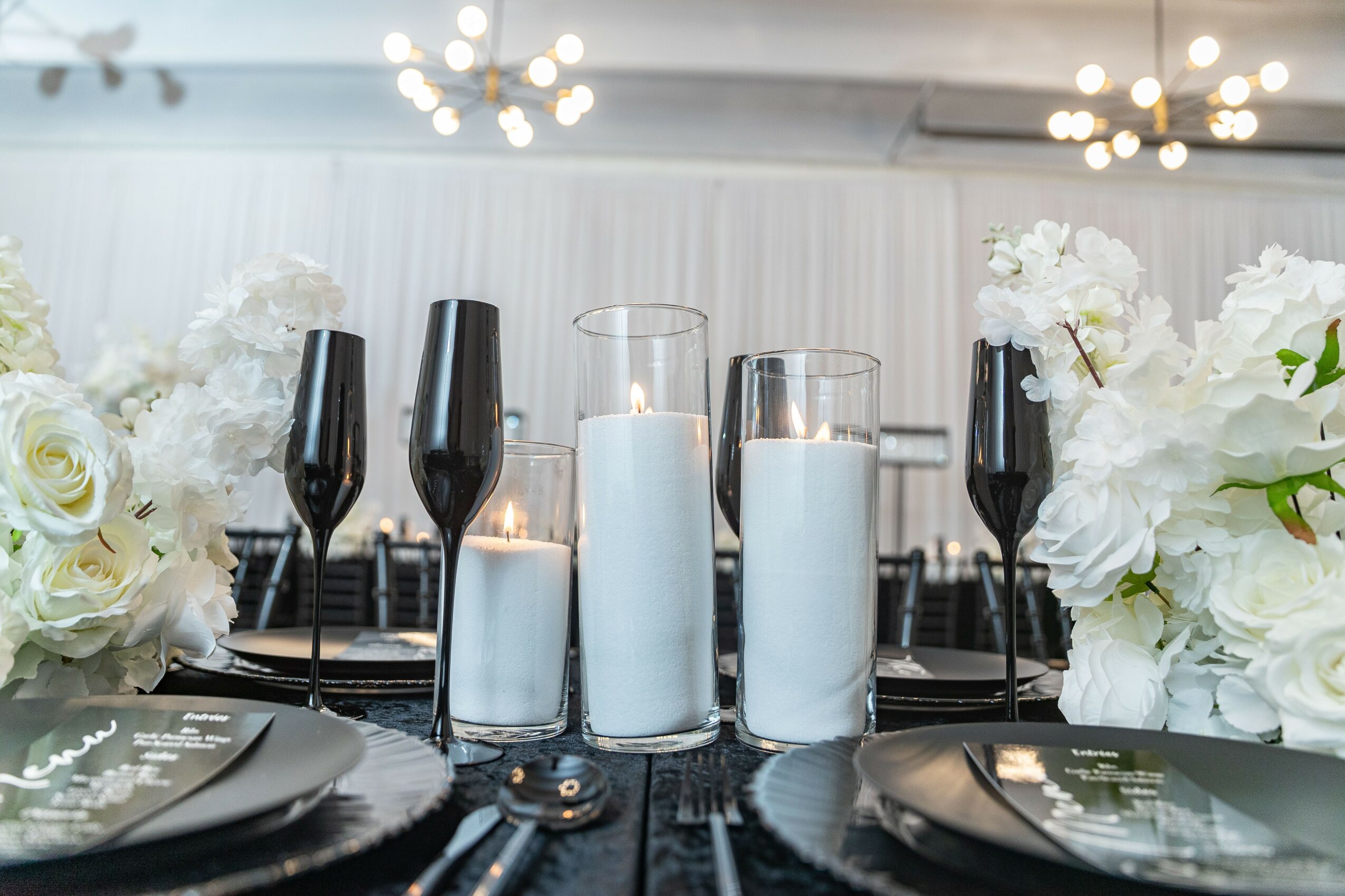 Table setting with black plates, tall glasses with white candles, and white floral arrangements, in a bright room.