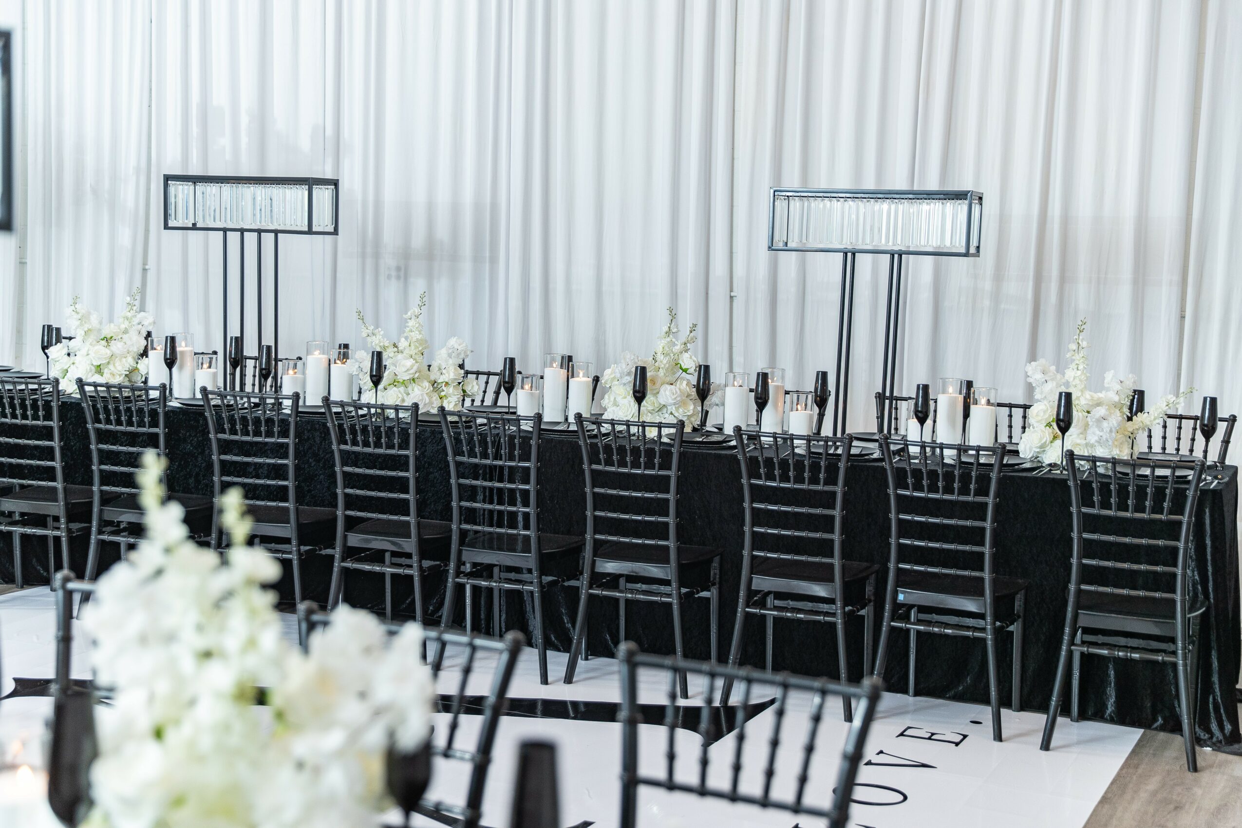Long banquet table with black chairs, white floral centerpieces, and tall empty frames, set against a white curtain backdrop.