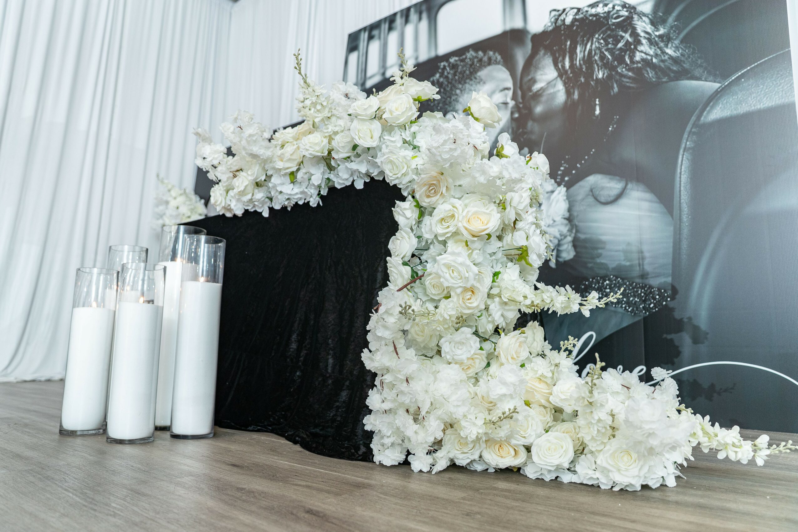 White floral arrangement on a black table with candles, against a black and white background, on a wooden floor.