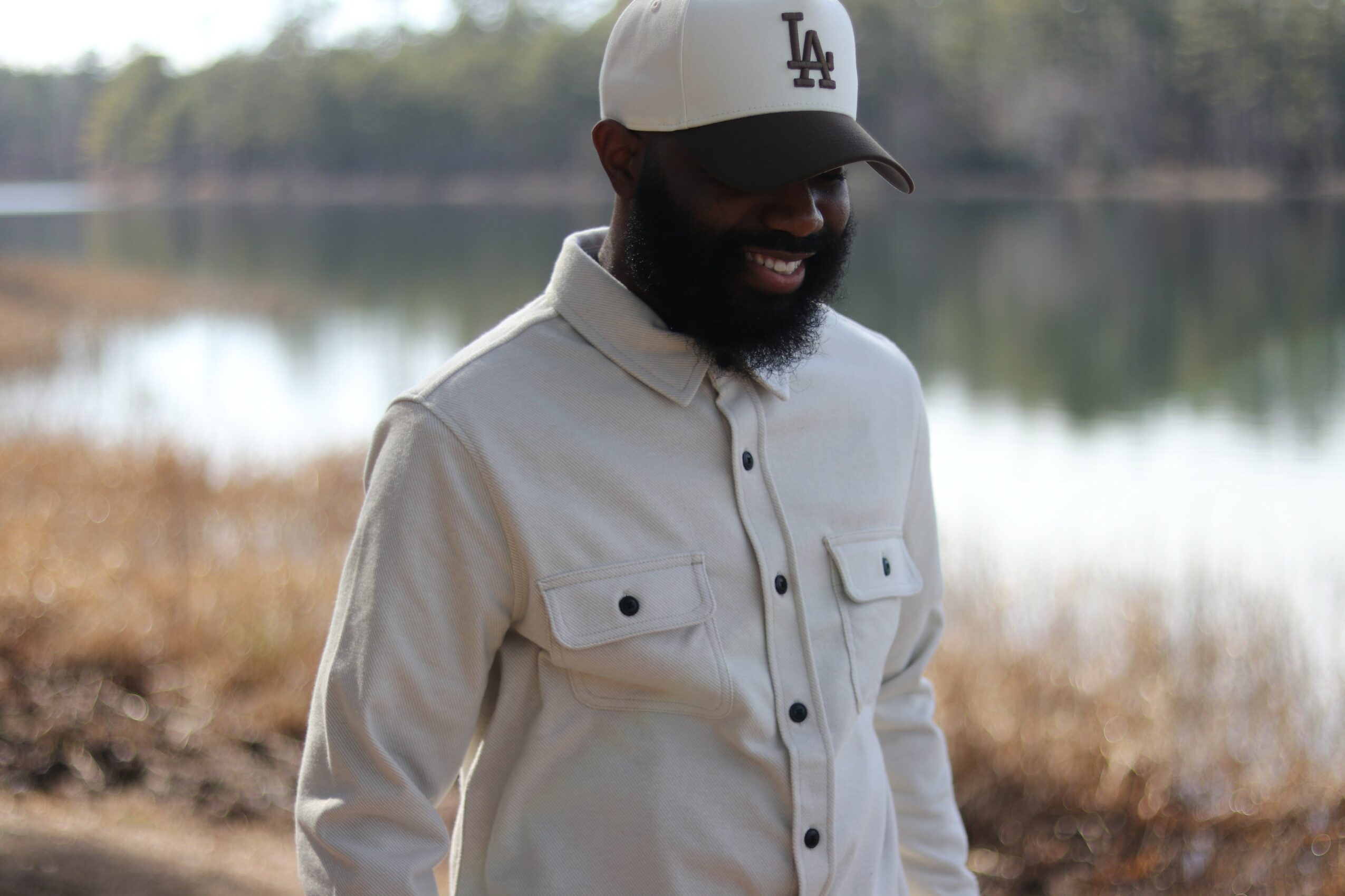 Man with a beard wearing a white cap and light-colored shirt outdoors near a lake, smiling.
