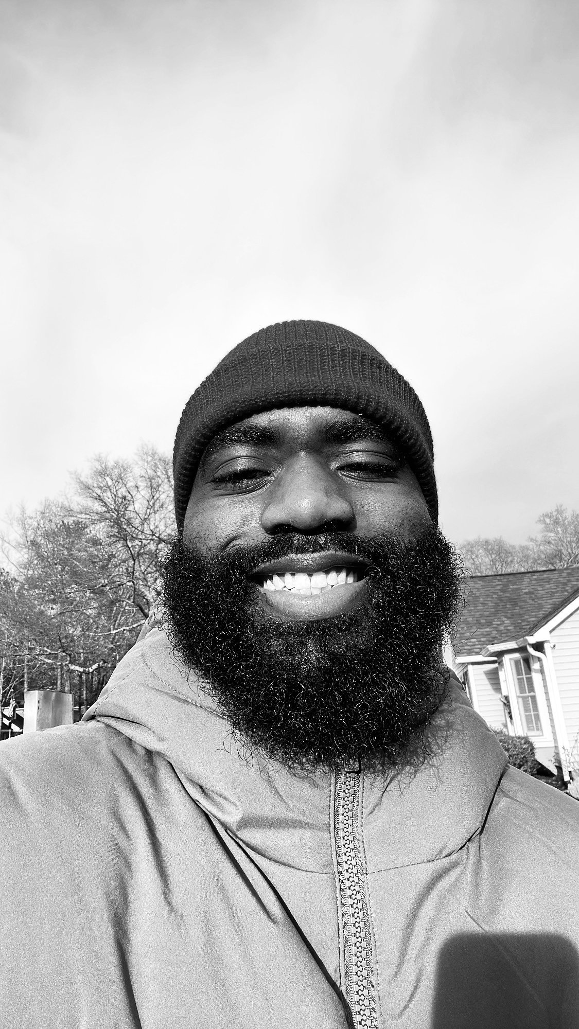 Smiling person with a beard wearing a beanie and jacket outdoors, houses and trees in background.