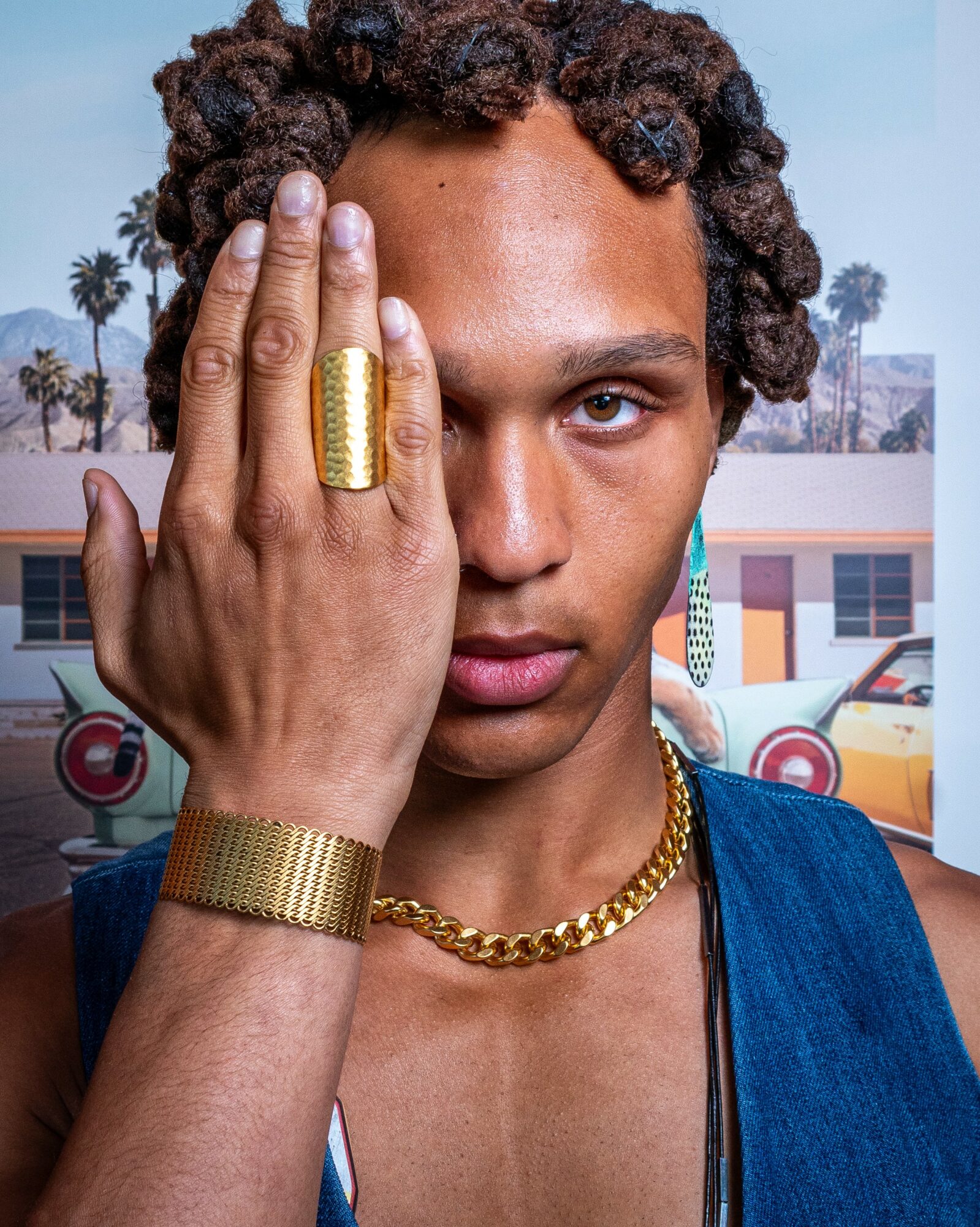 Young person with short curly hair covering one eye with hand, wearing gold jewelry, outdoors with palm trees and buildings.