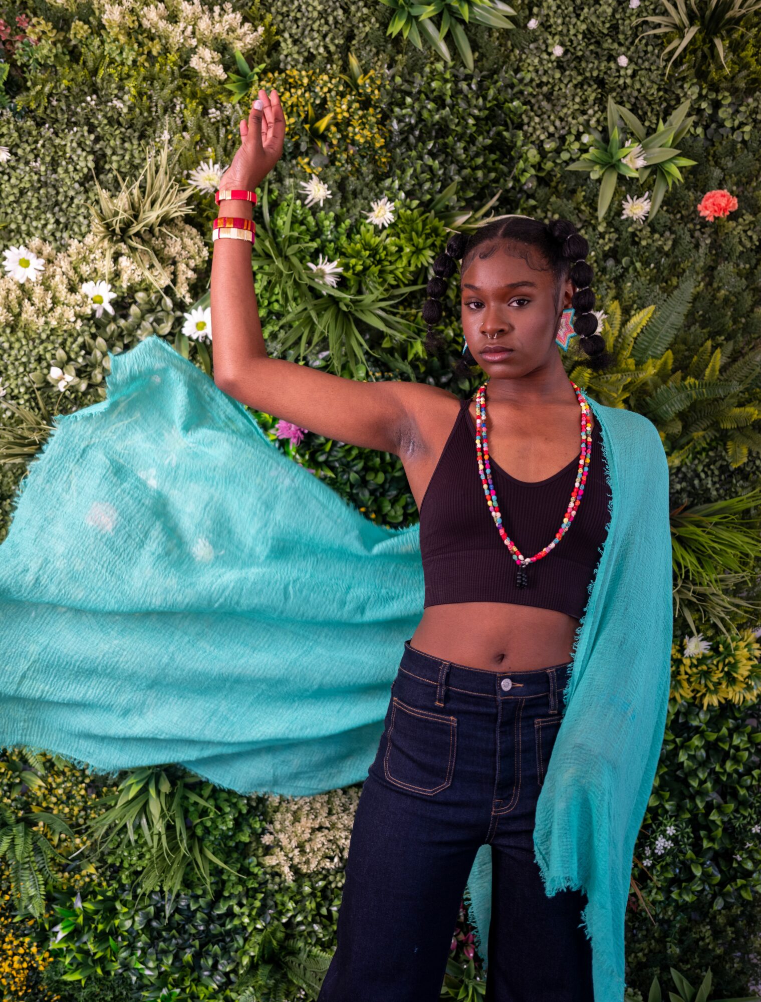 Young girl with braided hair, wearing a black crop top, jeans, and a turquoise shawl, standing against a floral background.