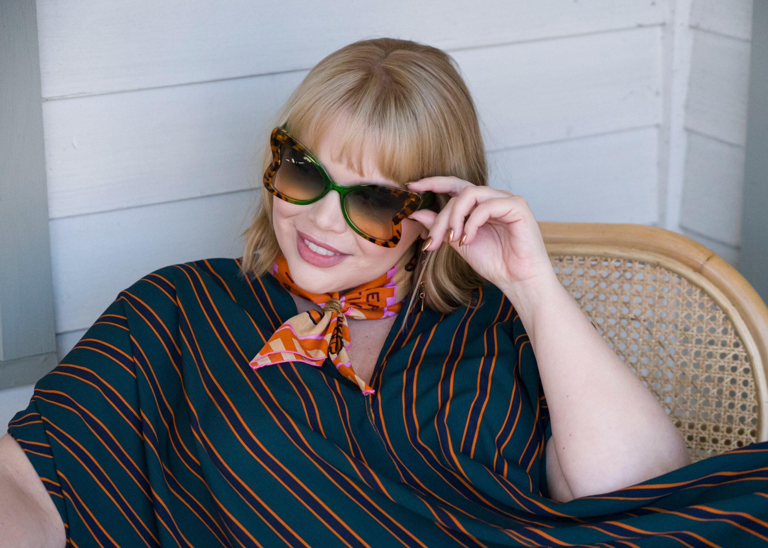 Woman with blonde hair wearing sunglasses, a striped shirt, and a colorful scarf, smiling and sitting on a chair.