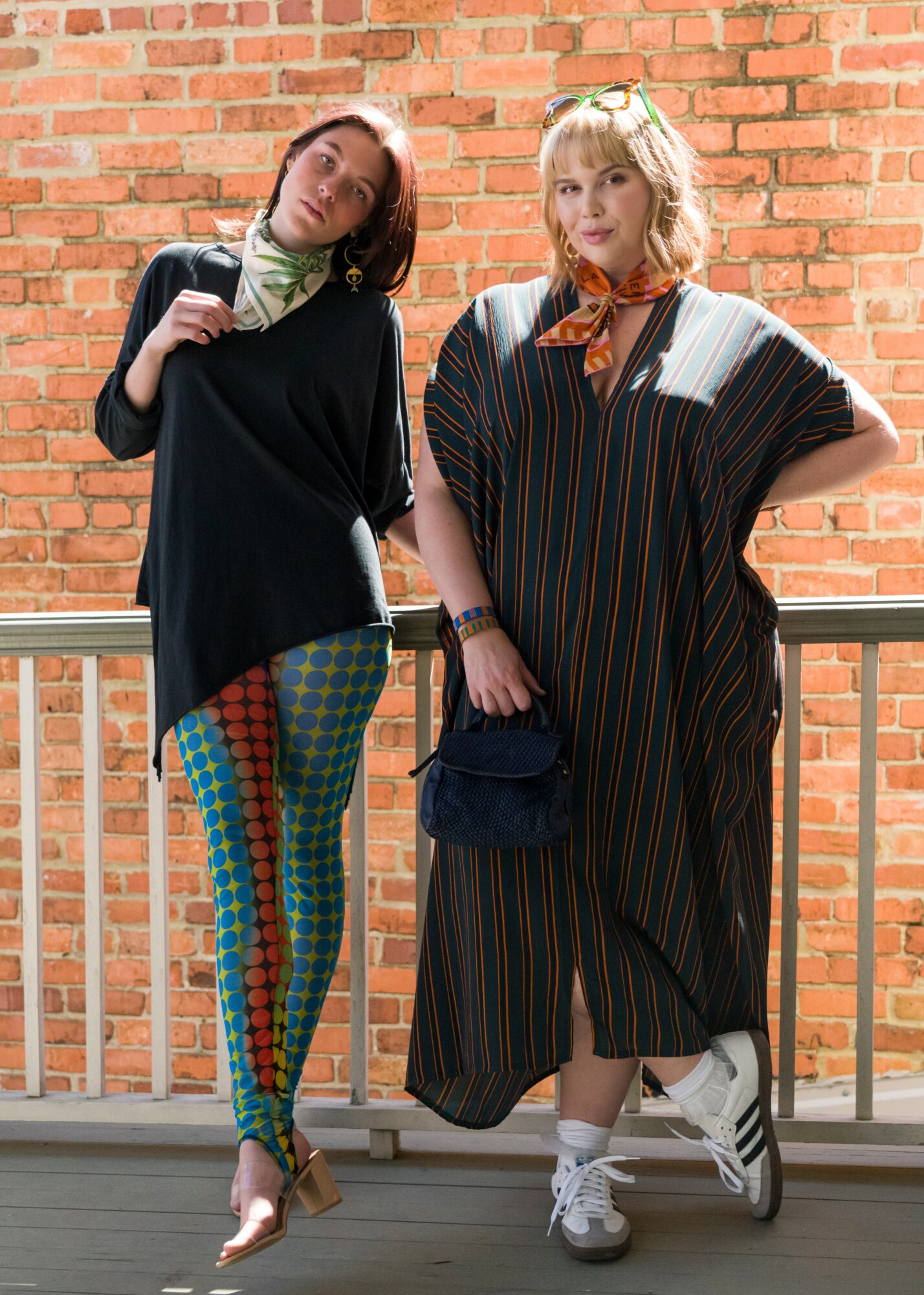 Two women standing on a balcony with a brick wall background, one in colorful patterned leggings and the other in a striped dress, both posing casually.