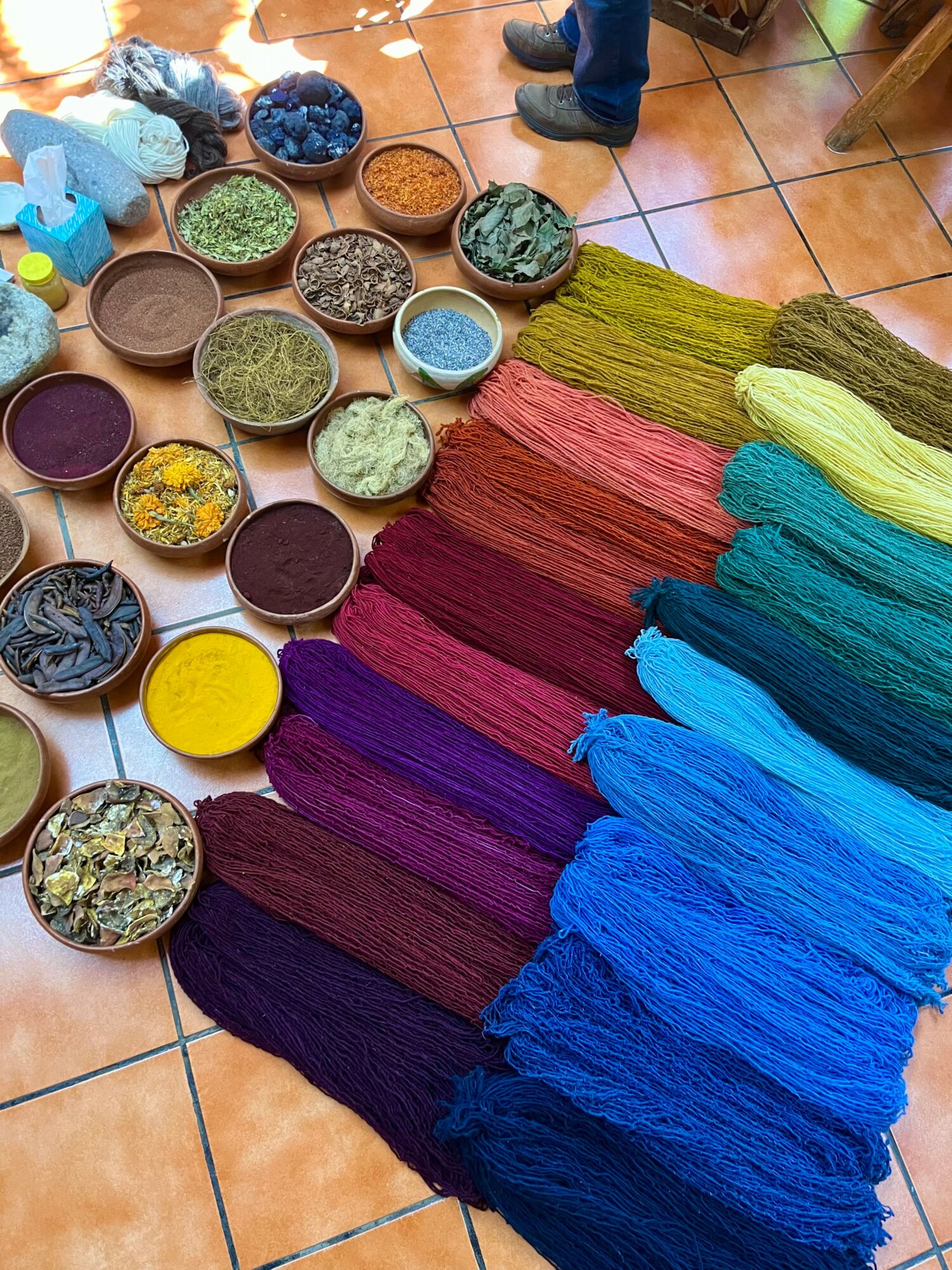 Colorful yarns arranged in rows with bowls of dyed fibers and materials on a tiled floor.