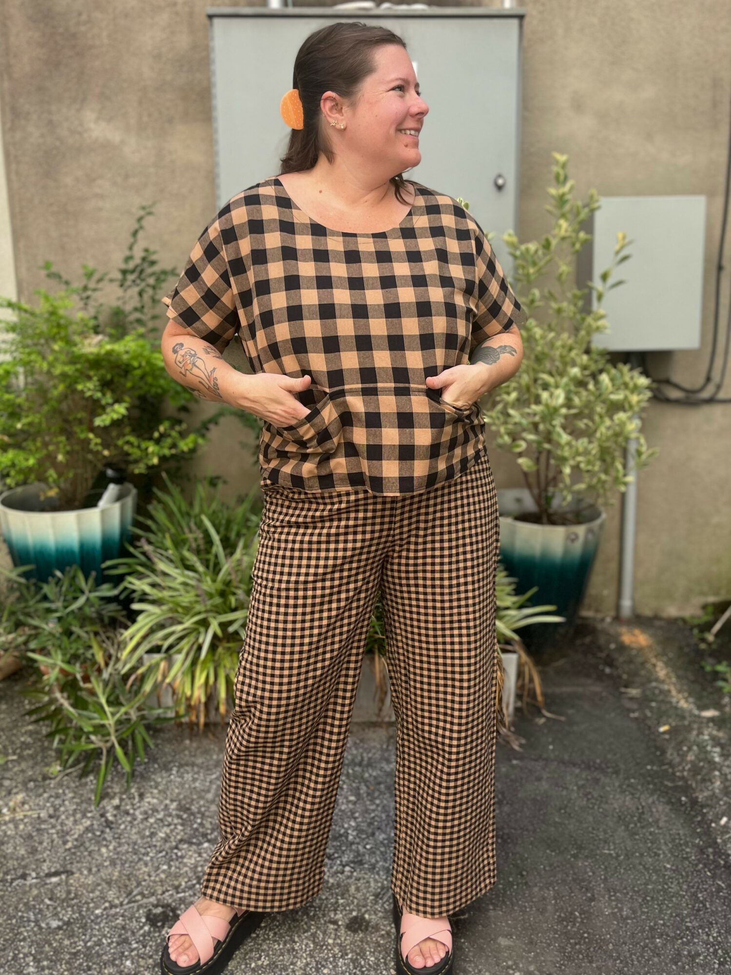 Woman standing outdoors with plants, smiling, wearing checkered top and pants, hands in pockets, facing right.